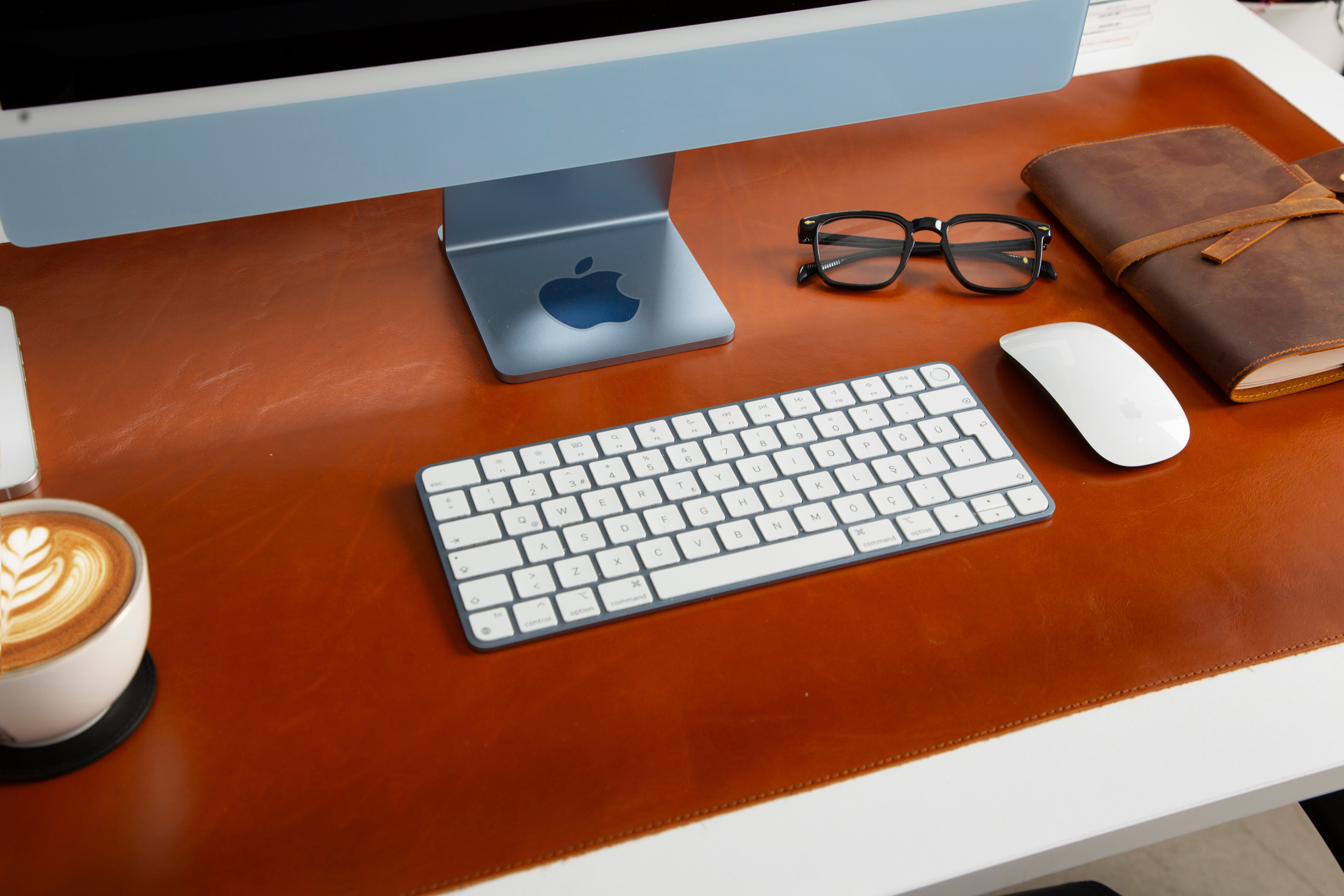 Bir Apple iMac, klavye, fare, gözlük, defter ve kahve masanın üzerinde.