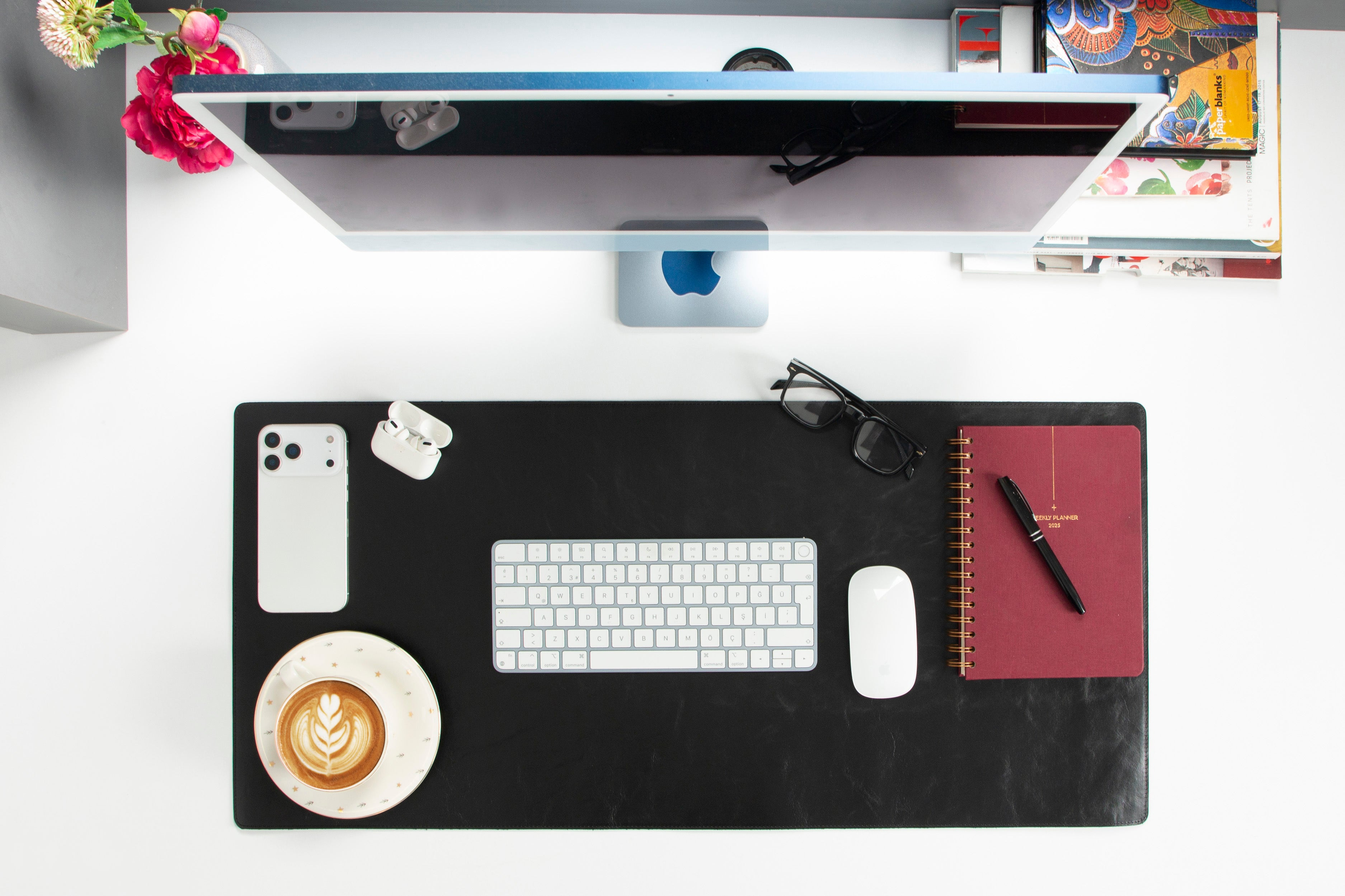Masa başında Apple monitör, klavye, fare, telefon, kahve, planlayıcı, gözlük ve çiçekler.