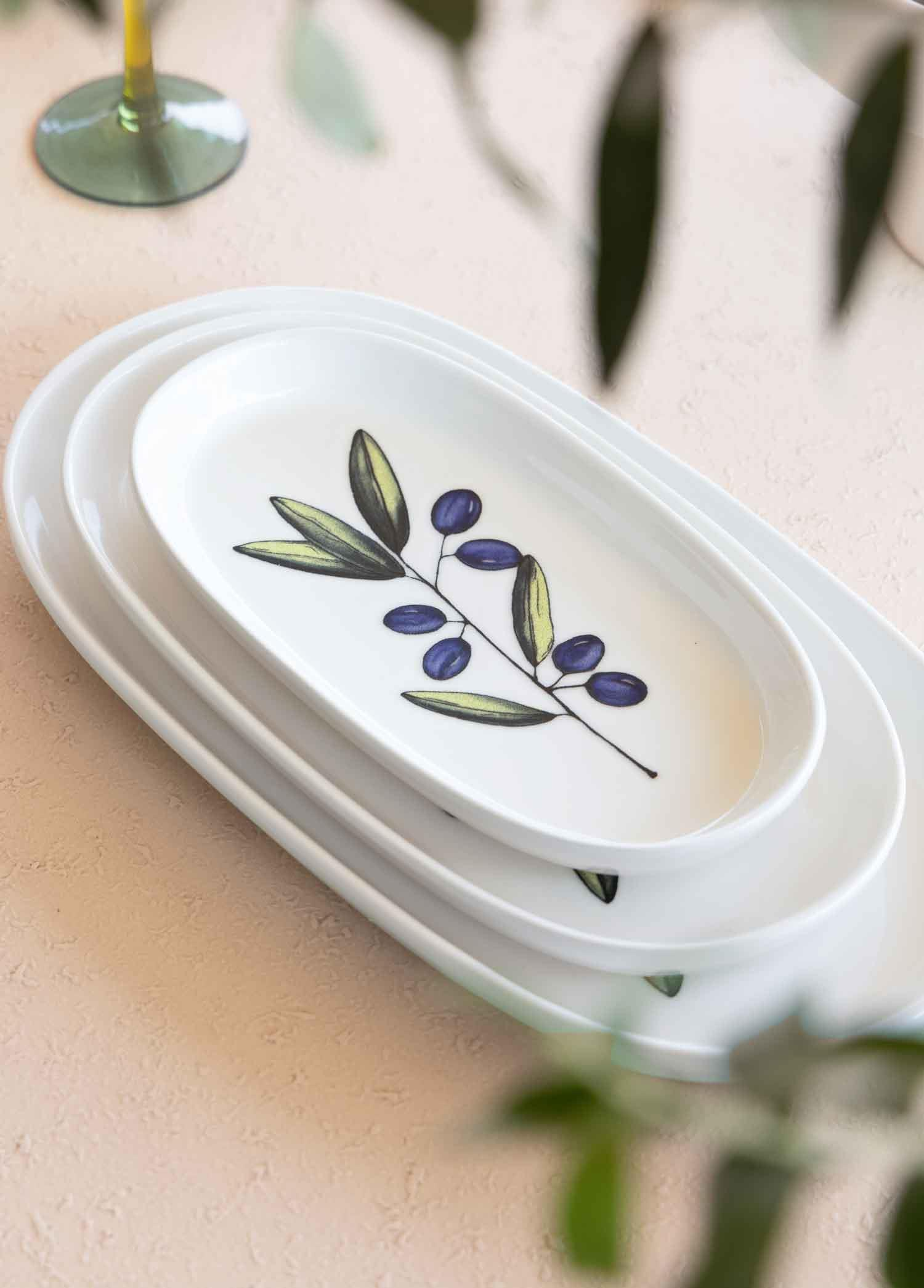 Pembe masa örtüsü üzerinde zeytin dalı desenli üç beyaz servis tabağı.