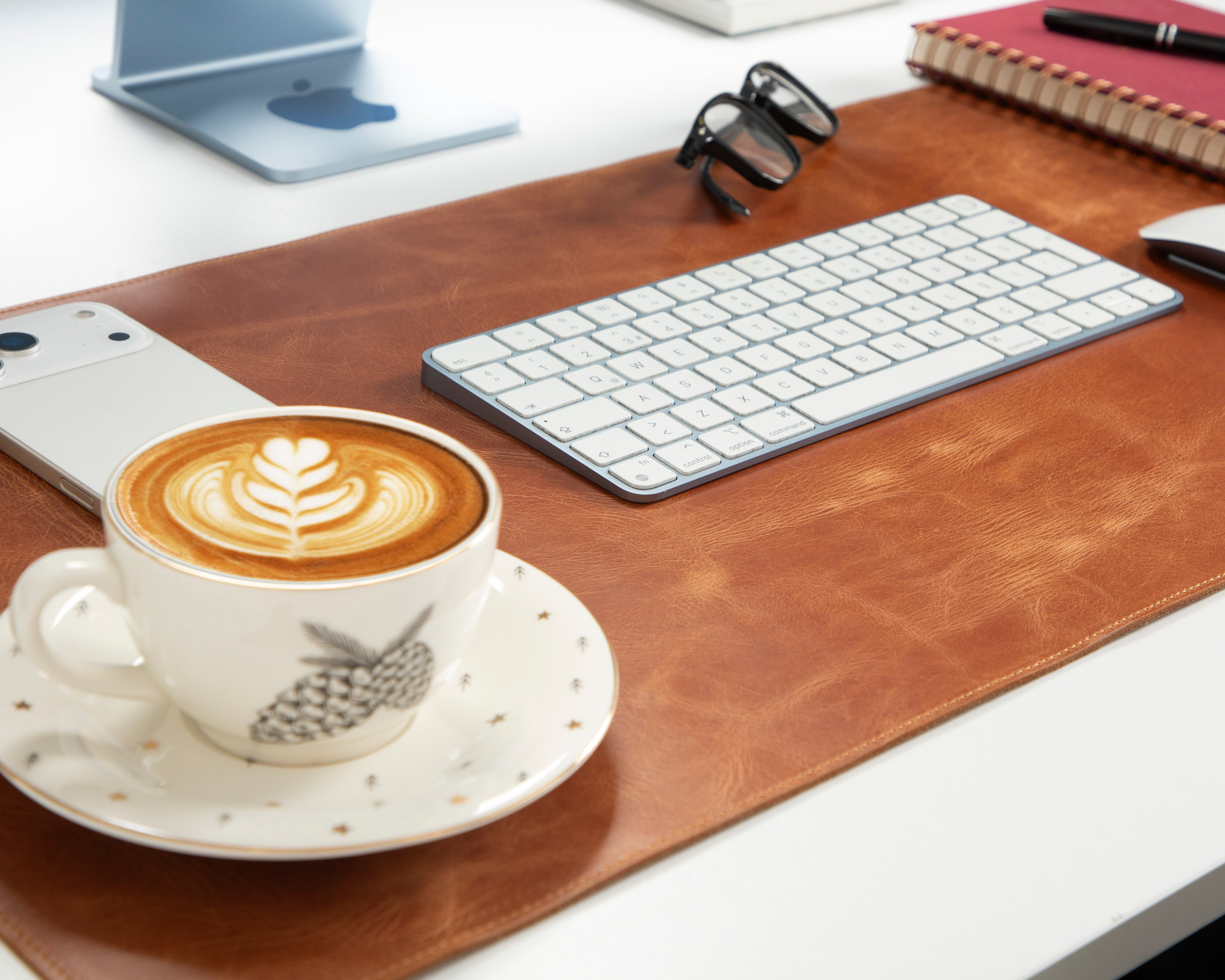 Masa üzerinde latte art'lı kahve fincanı, telefon, klavye, gözlük ve defter.