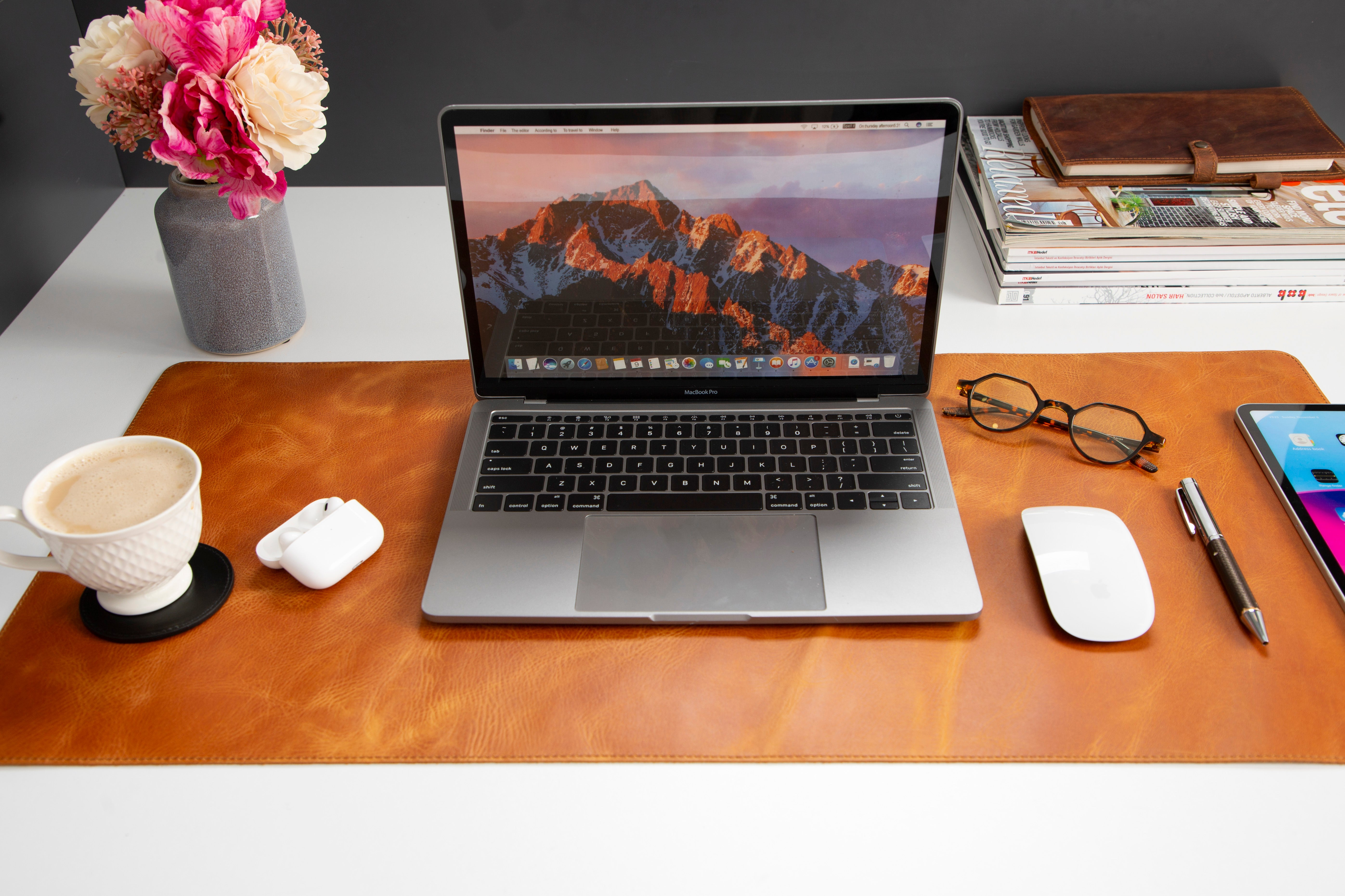 Laptop, fare, kahve, çiçek ve gözlük bulunan bir masa.