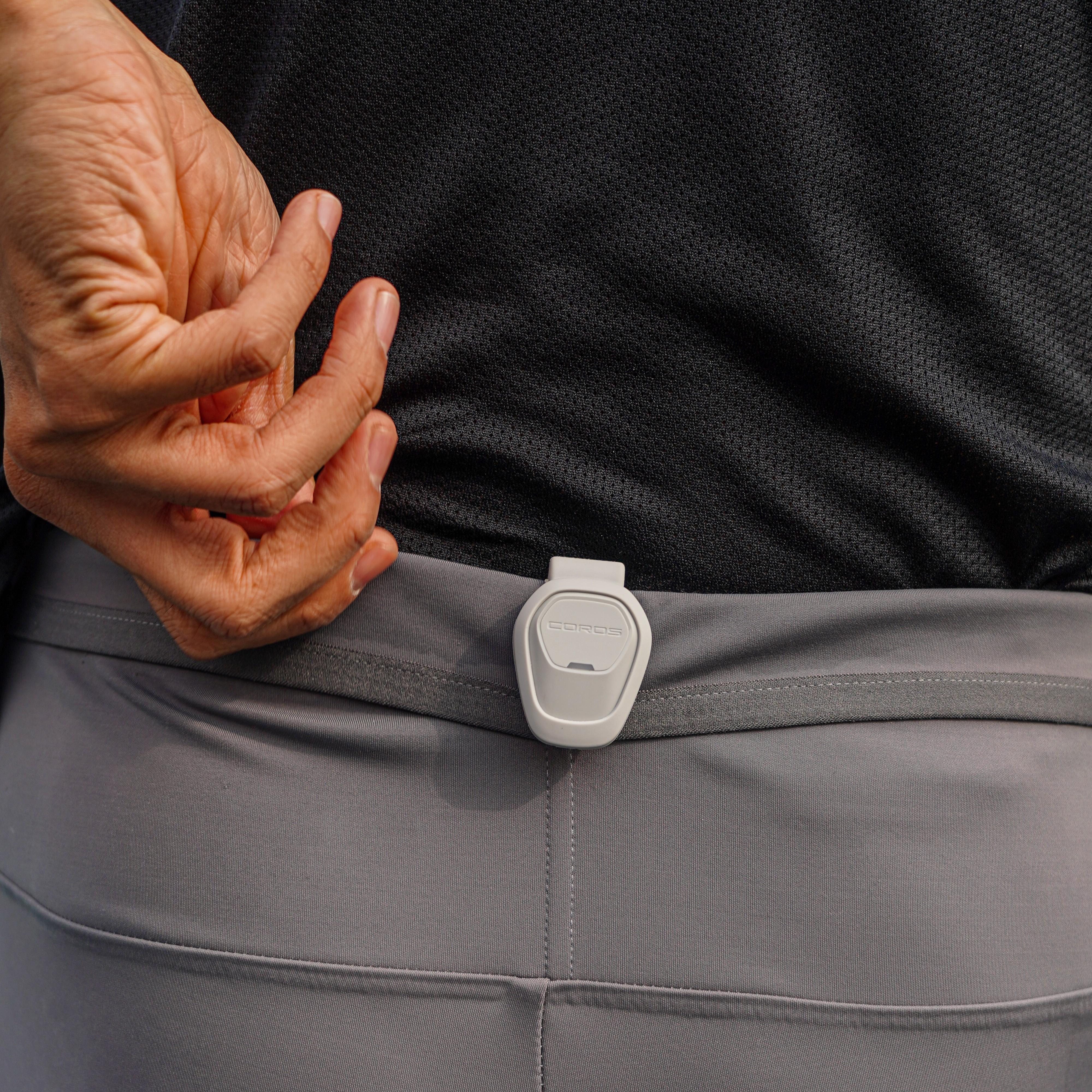 Una persona che indossa una maglietta nera e pantaloni grigi, con un dispositivo COROS bianco agganciato alla cintura posteriore.