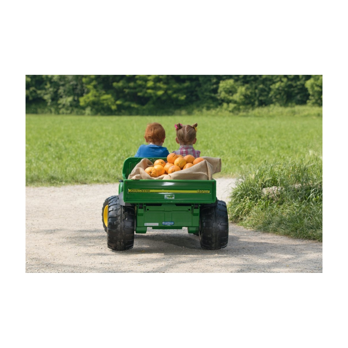 Zwei Kinder sitzen in einem grünen John Deere Gator mit Orangen im Laderaum.