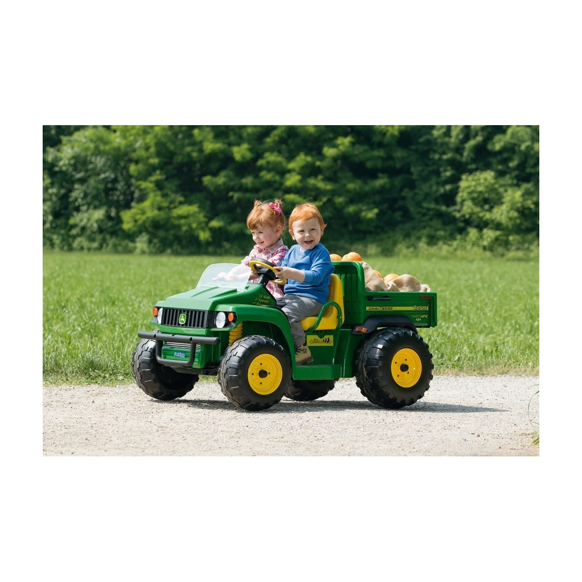 Zwei Kinder fahren in einem grünen John Deere Spielzeugauto durch einen Feldweg.