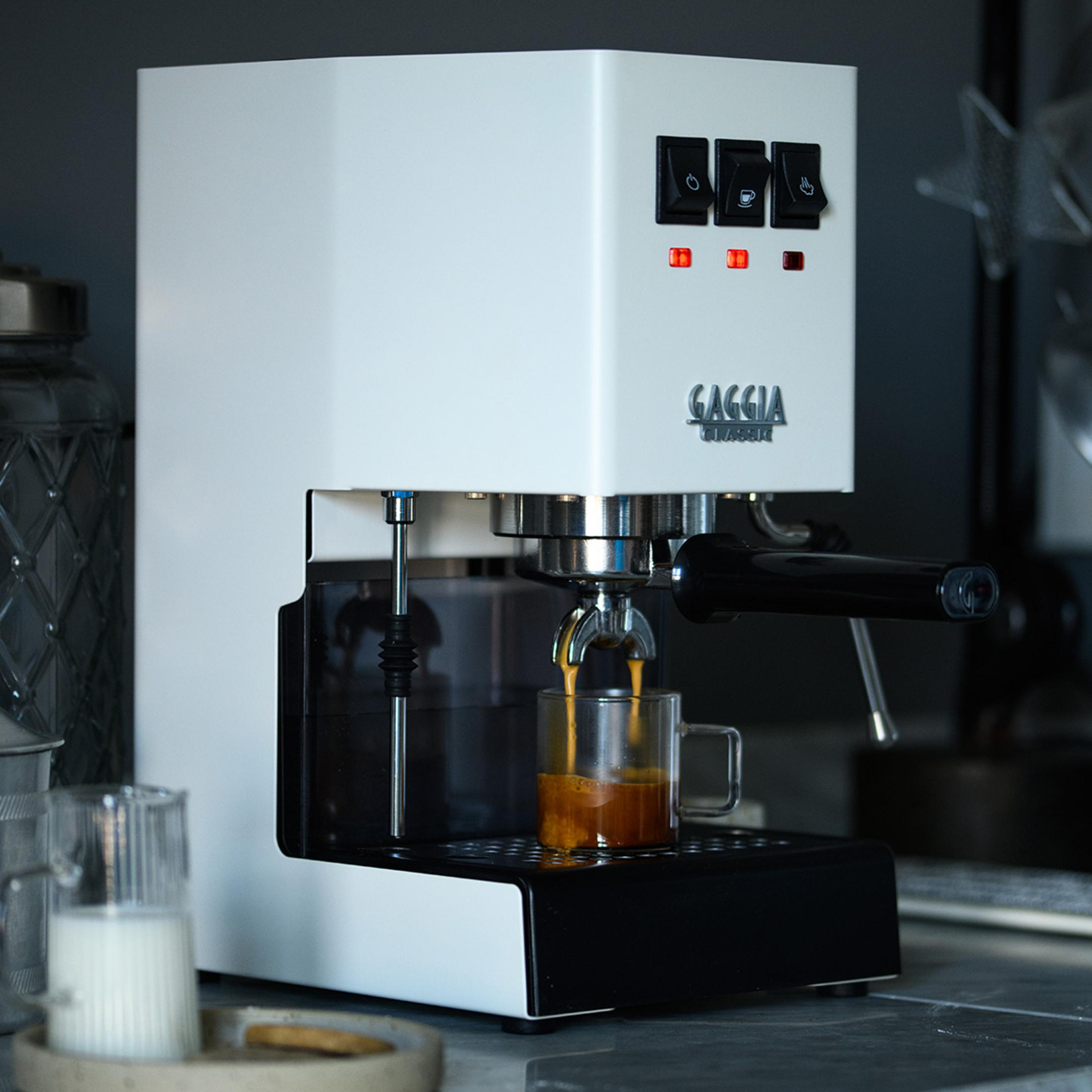 Een witte espressomachine zet koffie in een klein glas. Links staat een melkkannetje.