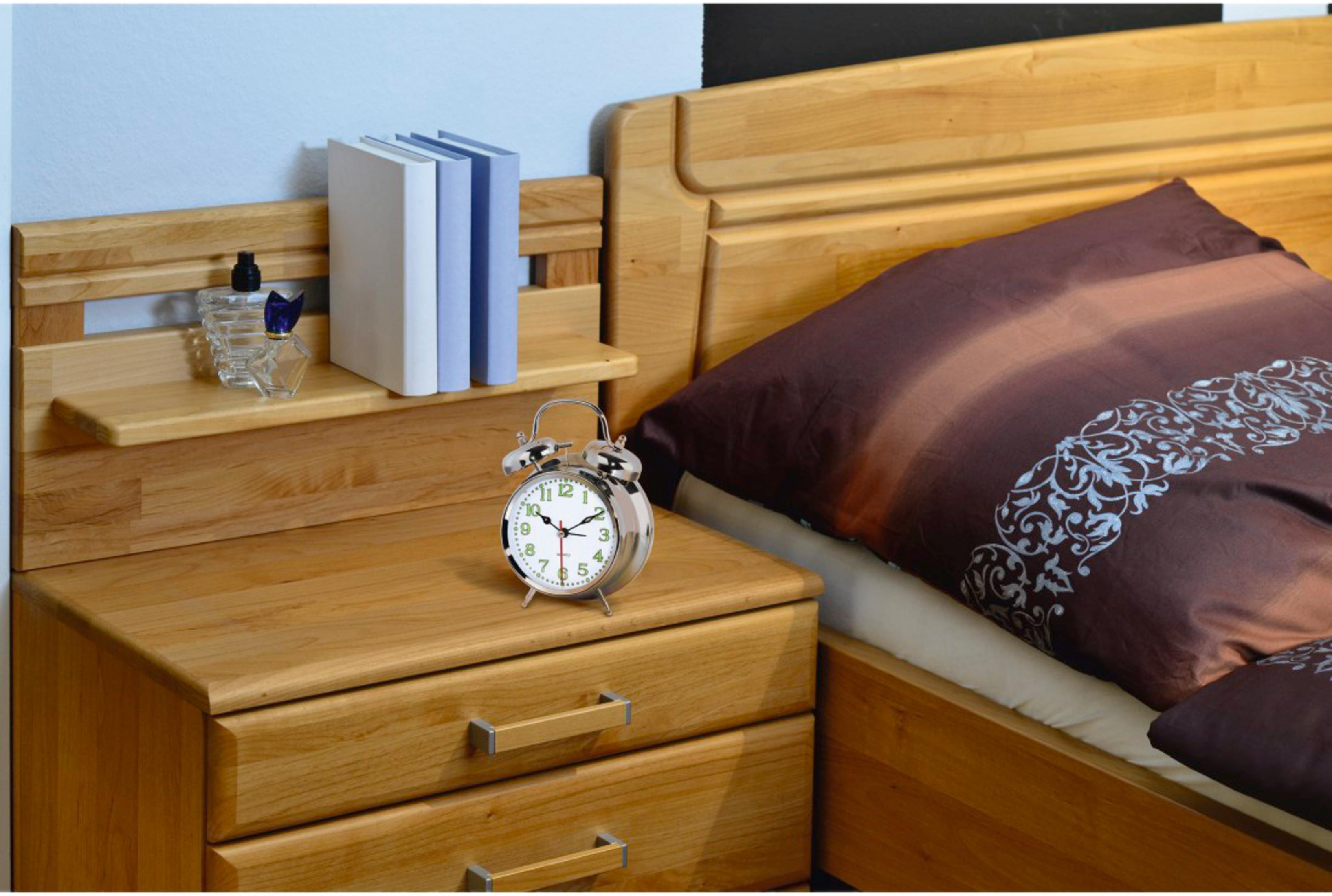Una mesita de noche de madera con despertador, libros y perfume junto a una cama con ropa de cama marrón.