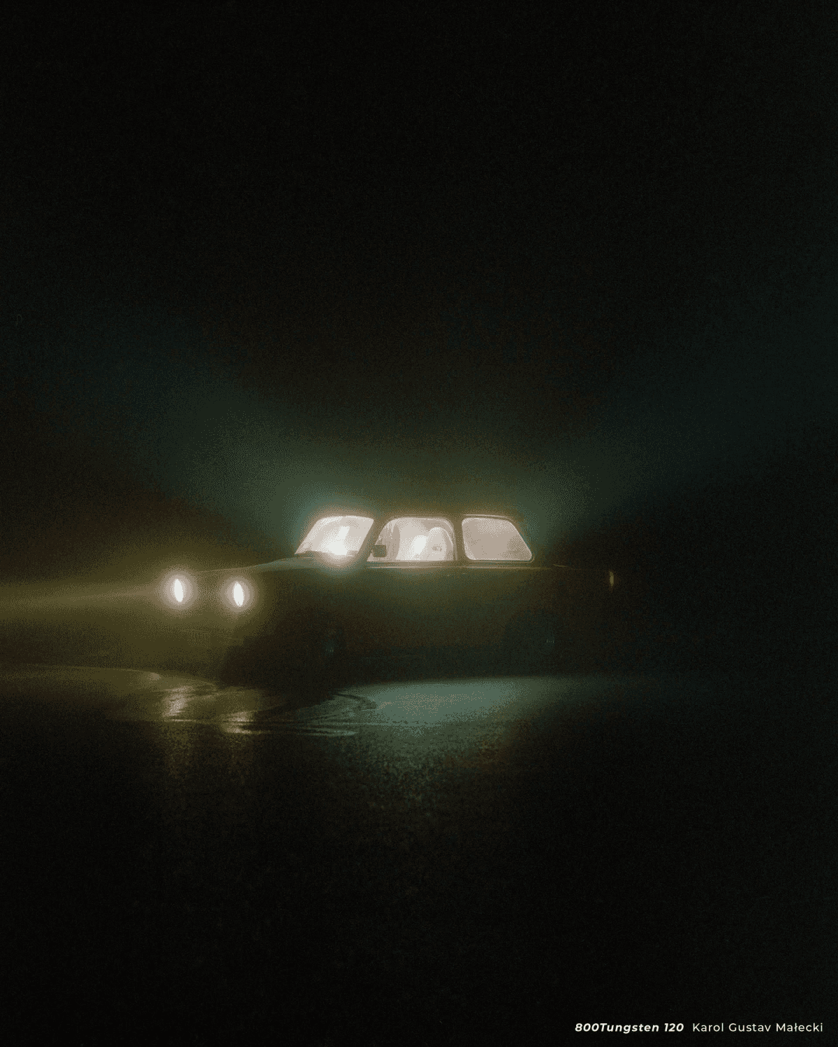 Un coche circula por una carretera mojada de noche, con los faros encendidos.