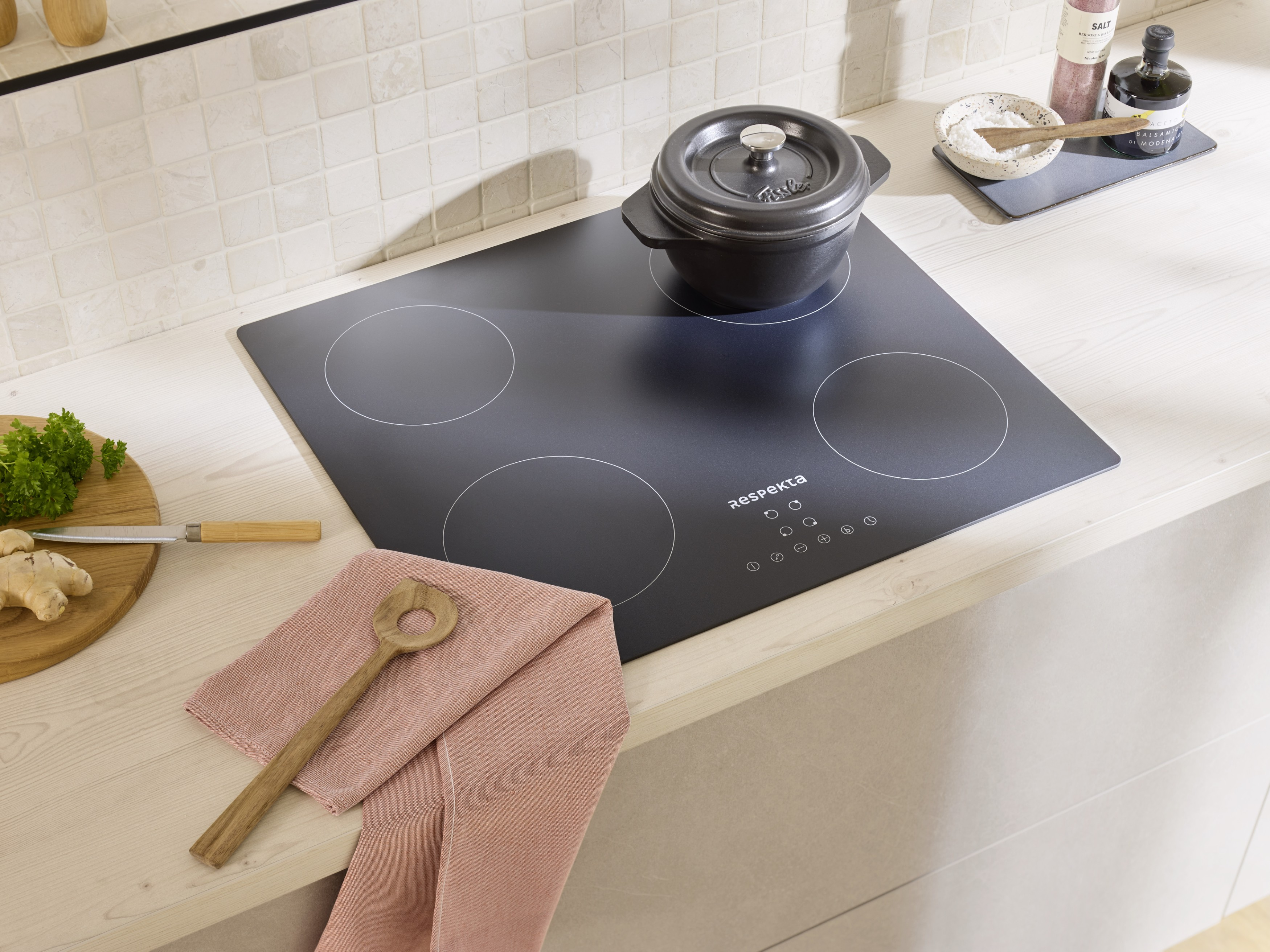 Une plaque à induction noire avec une cocotte, à côté une planche en bois avec du gingembre et du persil.