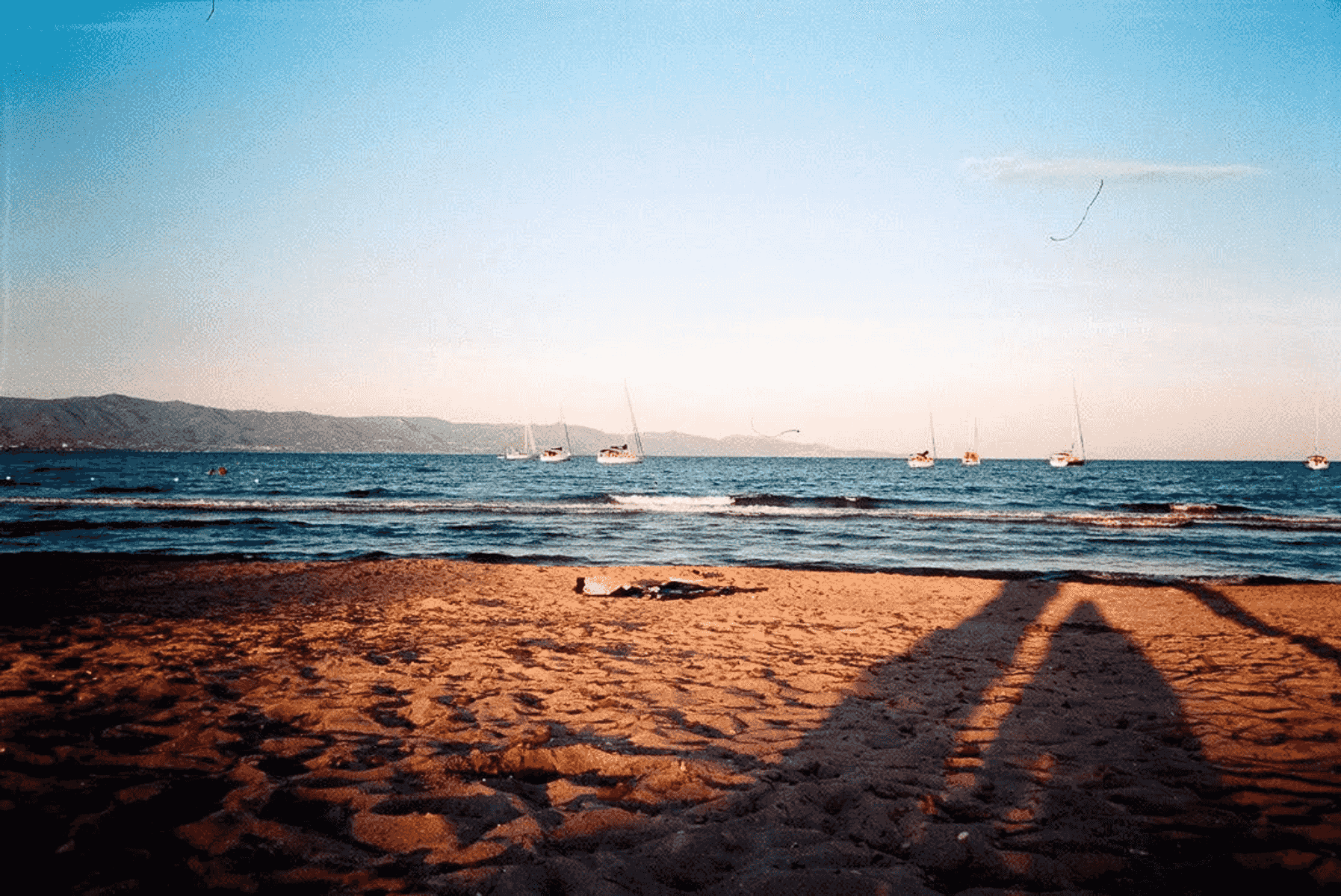 Una playa de arena con sombras, el mar y veleros en el horizonte.
