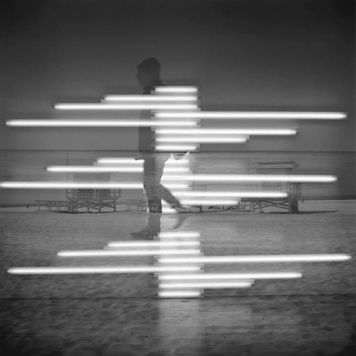 Una persona camina sobre arena, con luces de neón borrosas sobre una playa y torres de socorristas.