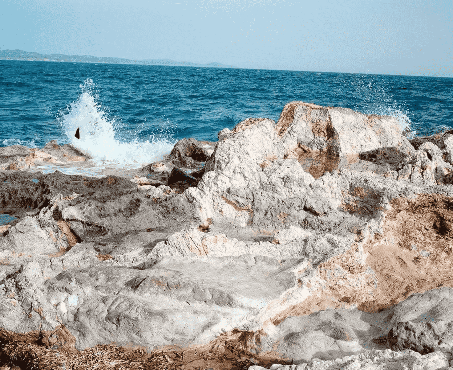 Una ola rompe en una costa rocosa con mar y cielo azules.