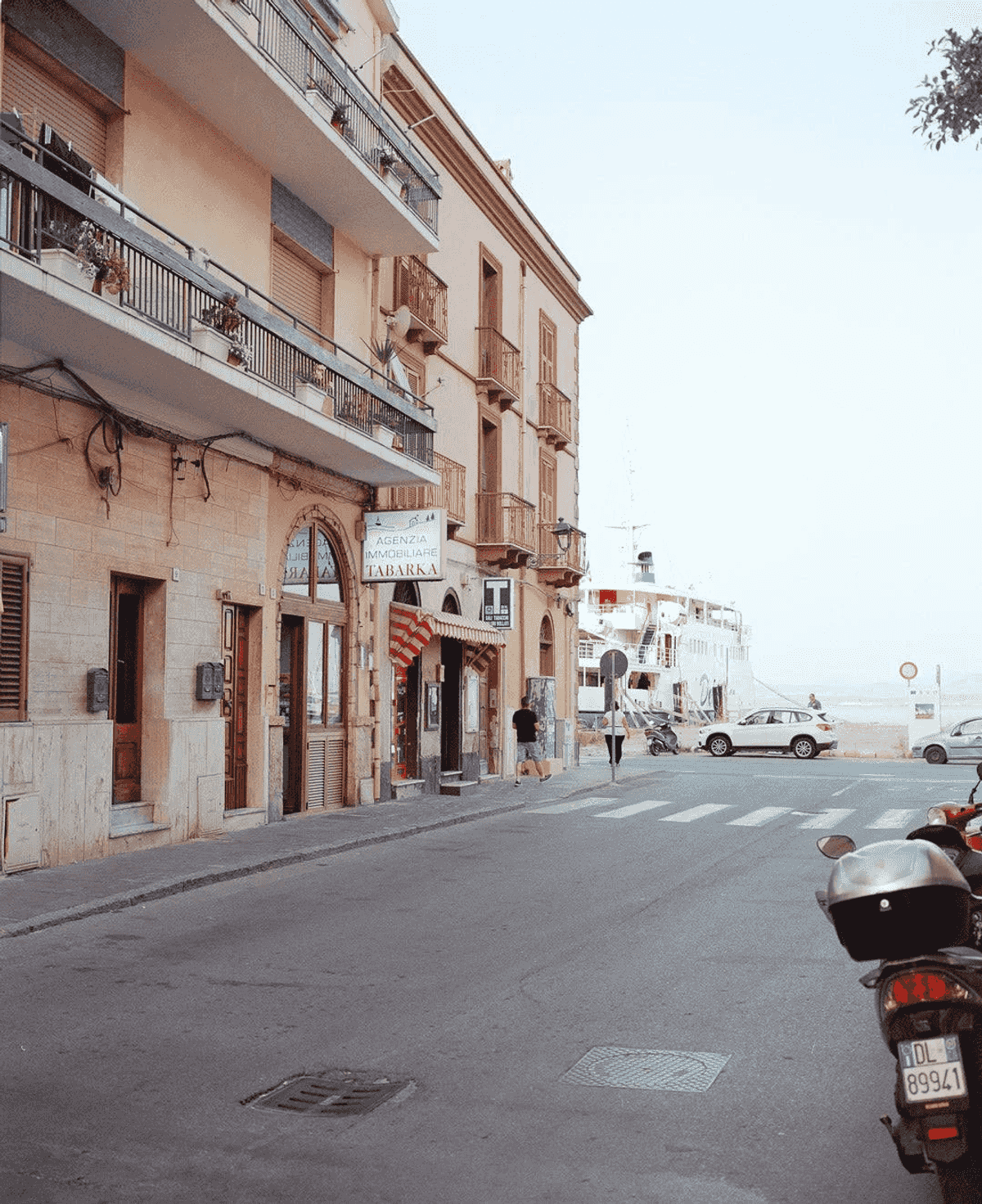 Edificios con balcones y tiendas, calle con coches, motos y gente.