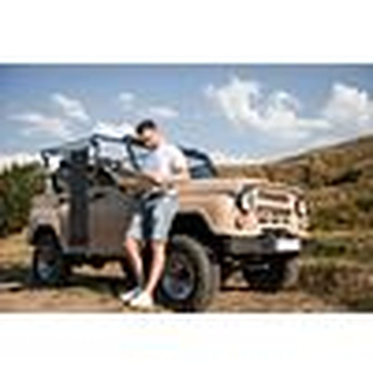 Un uomo in pantaloncini di jeans e maglietta bianca si appoggia a una Jeep beige fuoristrada.