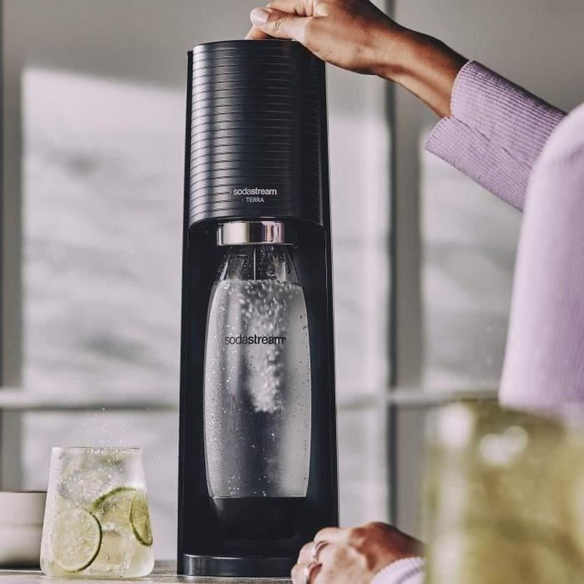 Osoba używa czarnej maszyny SodaStream Terra do nasycania wody w butelce. Obok stoi szklanka wody z plasterkami limonki.