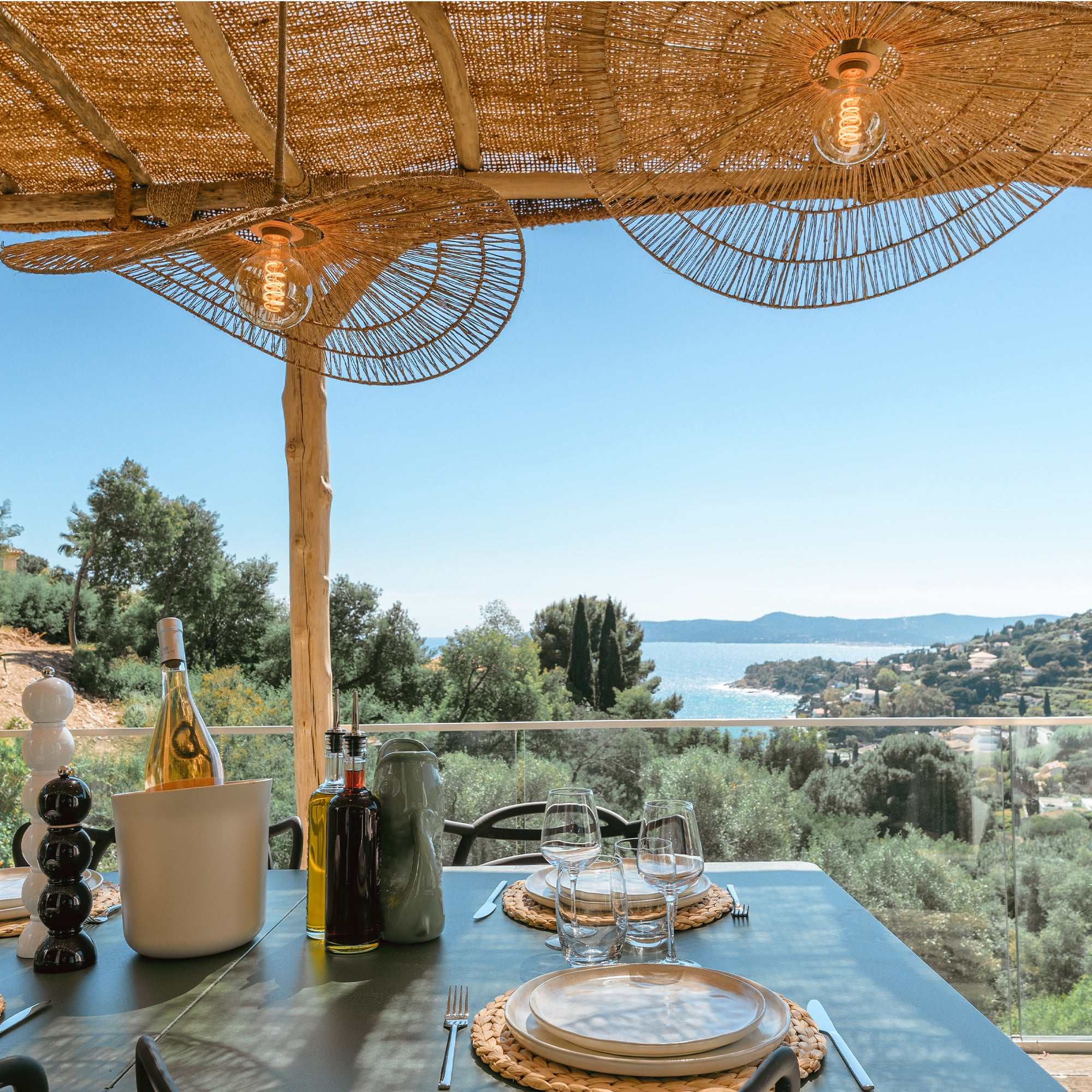 Gedeckter Tisch mit Meerblick, Sonnenschirm aus Stroh, Lampen und Flaschen.