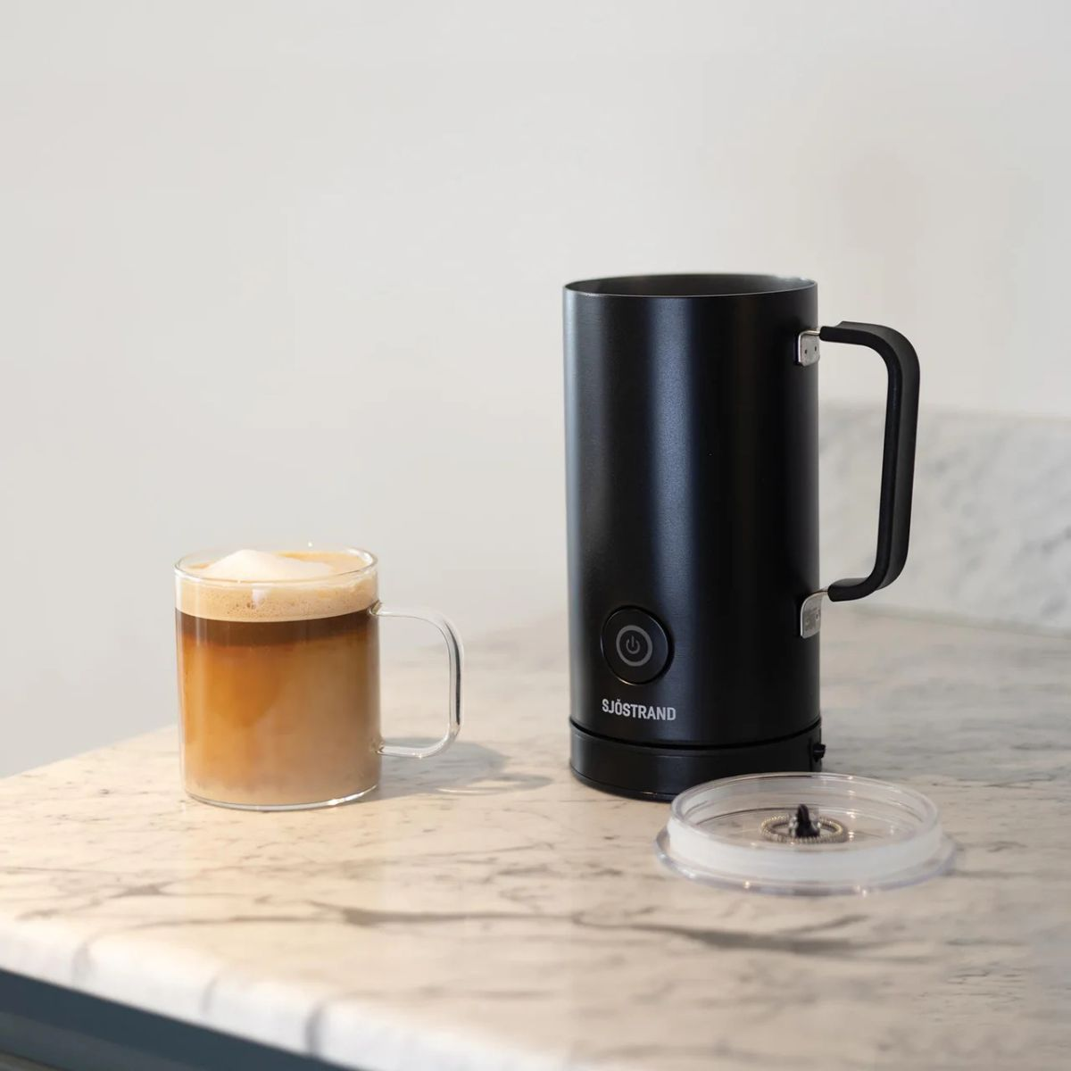 Ein Glas Latte Macchiato neben einem schwarzen Milchaufschäumer auf Marmorplatte.
