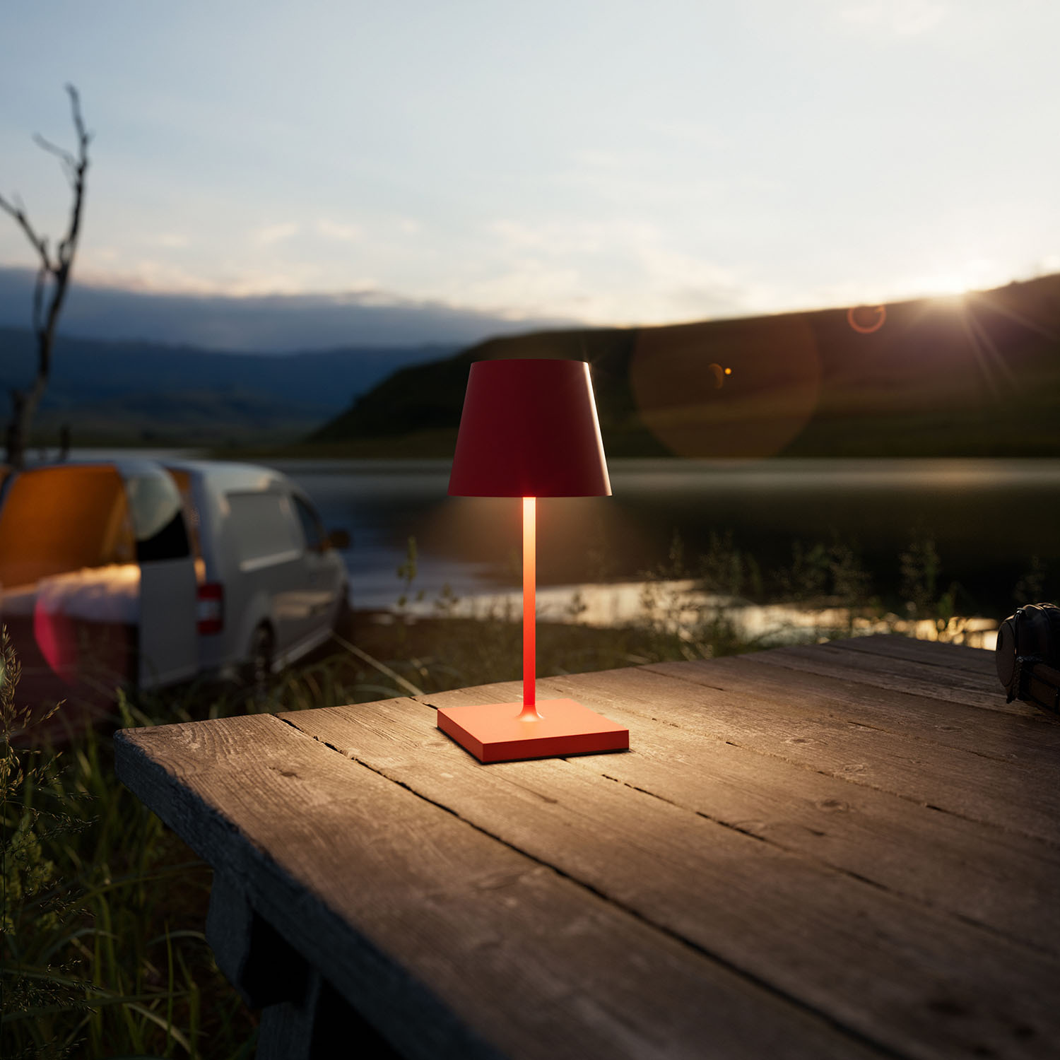 Eine rote Lampe beleuchtet einen Holztisch in der Dämmerung. Ein Wohnmobil und Zelte sind zu sehen.