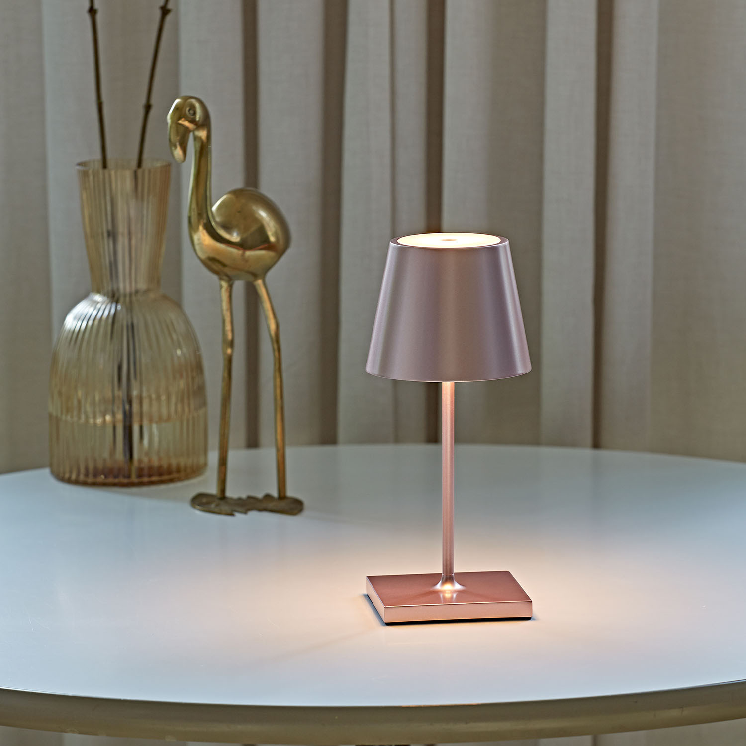 Eine Tischlampe aus Roségold steht auf einem weißen Tisch neben einer Vase mit Zweigen und einer Flamingo-Figur.