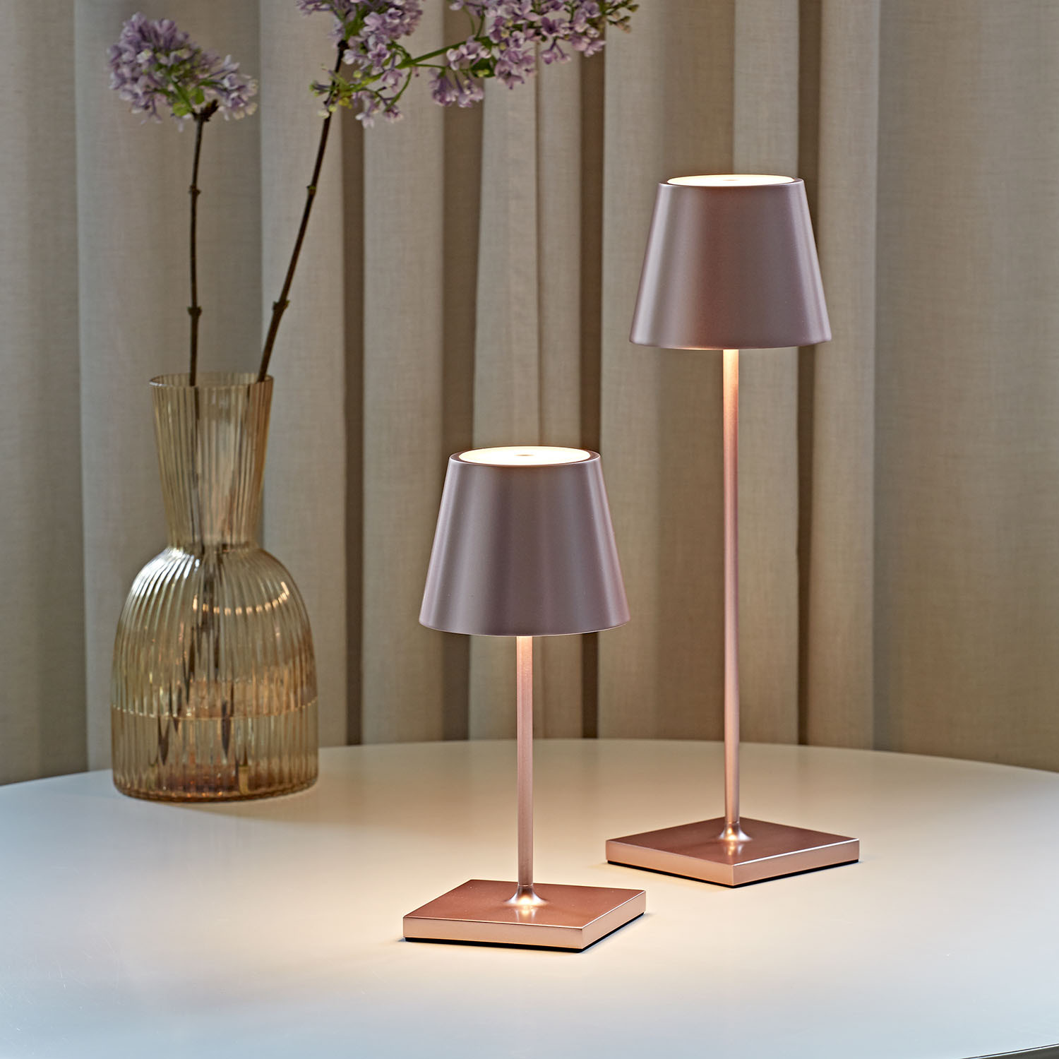 Zwei kupferfarbene Tischlampen mit rosa Schirmen stehen auf einem Tisch. Links eine Vase mit lila Blumen.