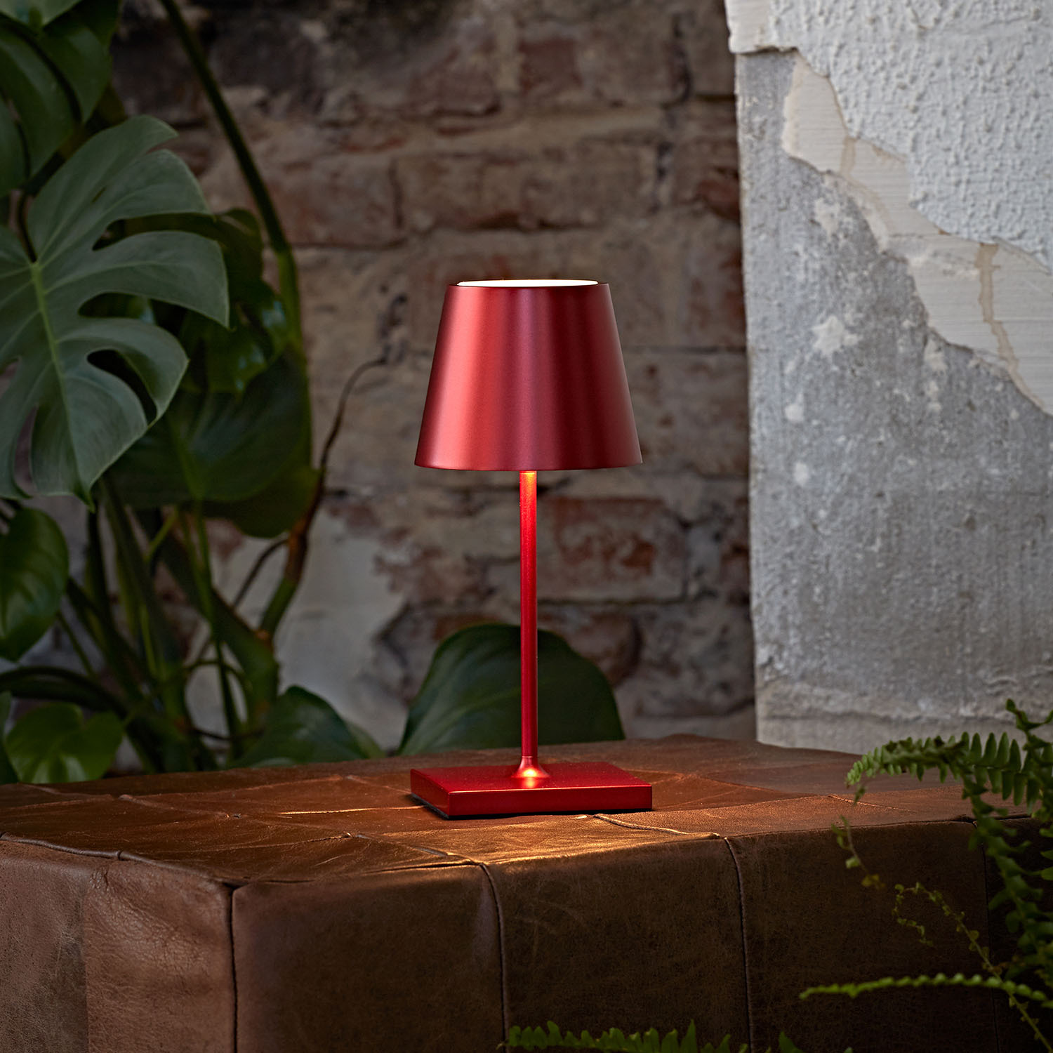 Eine rote kabellose Lampe steht auf einem braunen Lederhocker, mit Pflanzen und Ziegelwand im Hintergrund.