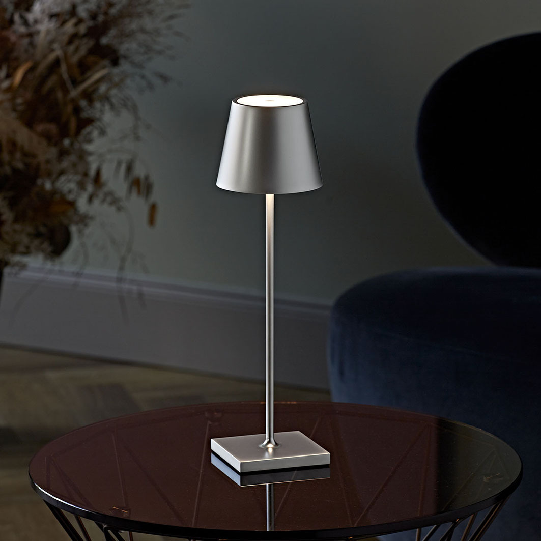 Eine schlanke silberne Tischlampe mit einem kegelförmigen Lampenschirm steht auf einem runden Tisch.