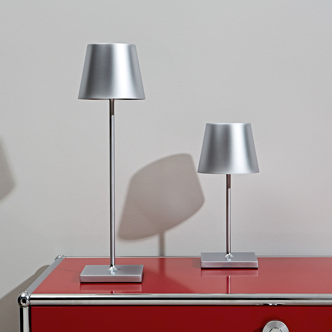 Zwei silberne Tischlampen mit schmalen Ständern auf einem roten Schrank vor einer grauen Wand.