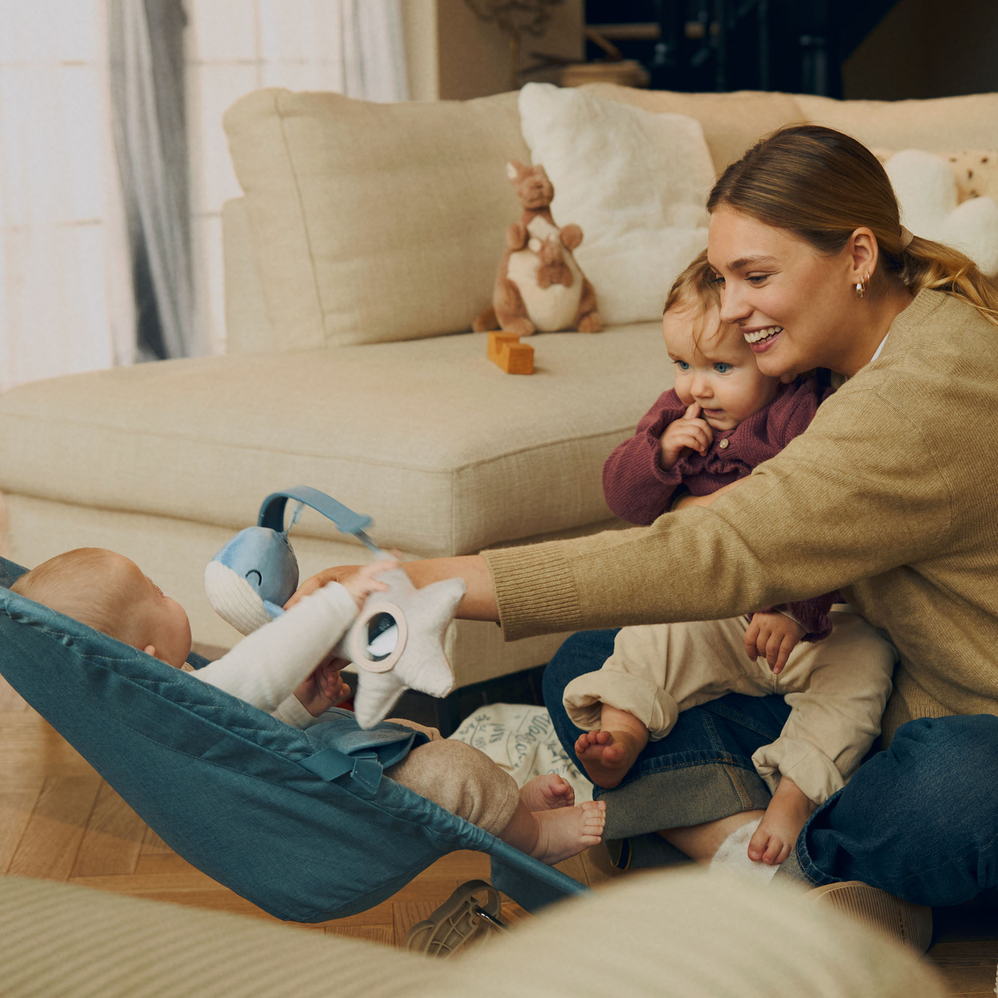 Eine Frau sitzt auf dem Boden und hält ein Baby. Ein anderes Baby liegt in einer blauen Hängematte.