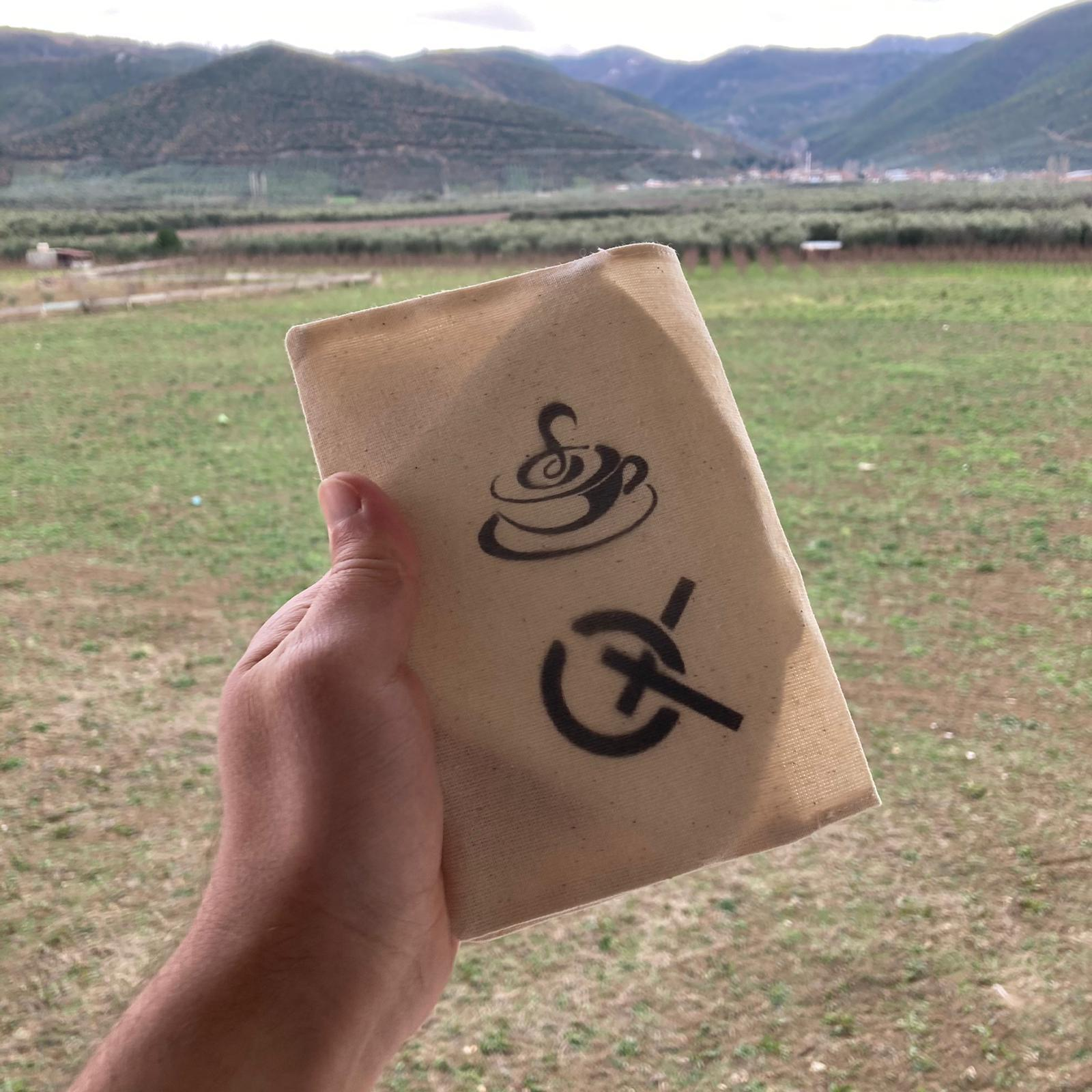 El, üzerinde kahve fincanı ve üzeri çizili sembol bulunan bej kumaşı tutuyor. Dışarıda dağlar ve tarlalar.