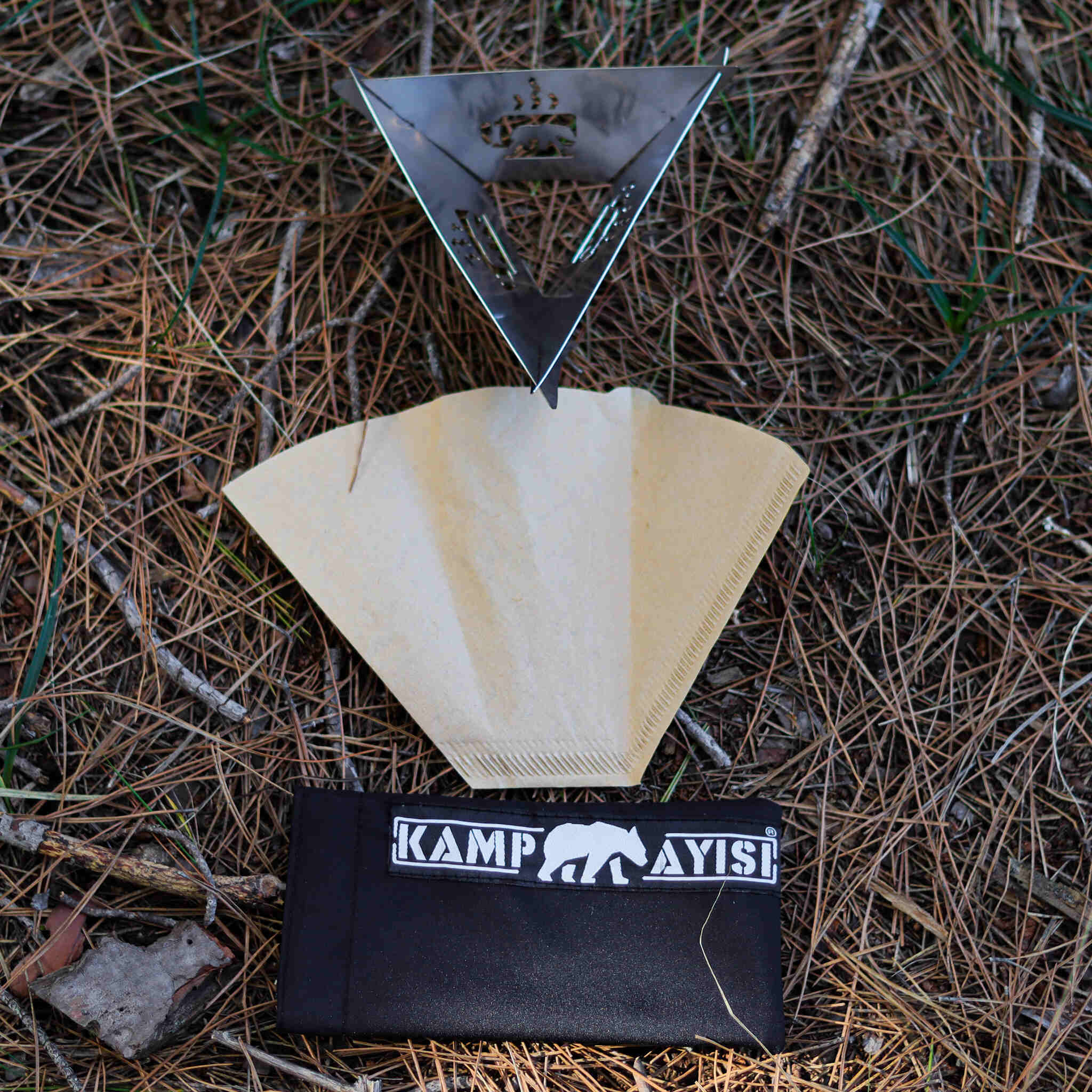Metal kamp ocağı, kahve filtresi ve siyah kese çam iğneleri üzerinde.