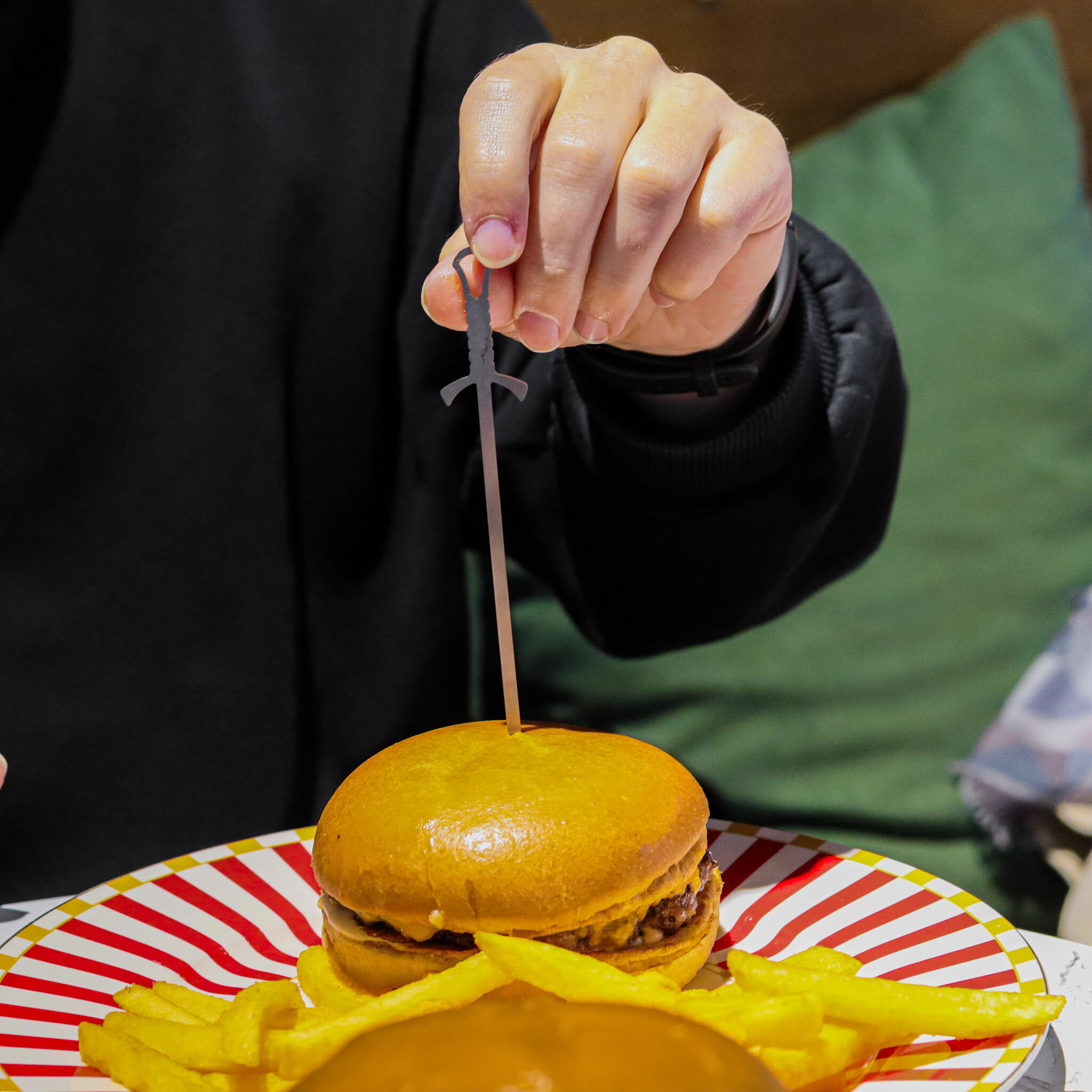 Bir elin tuttuğu, üzerinde kılıç şeklinde kürdan olan, patates kızartmalı burger tabağı.