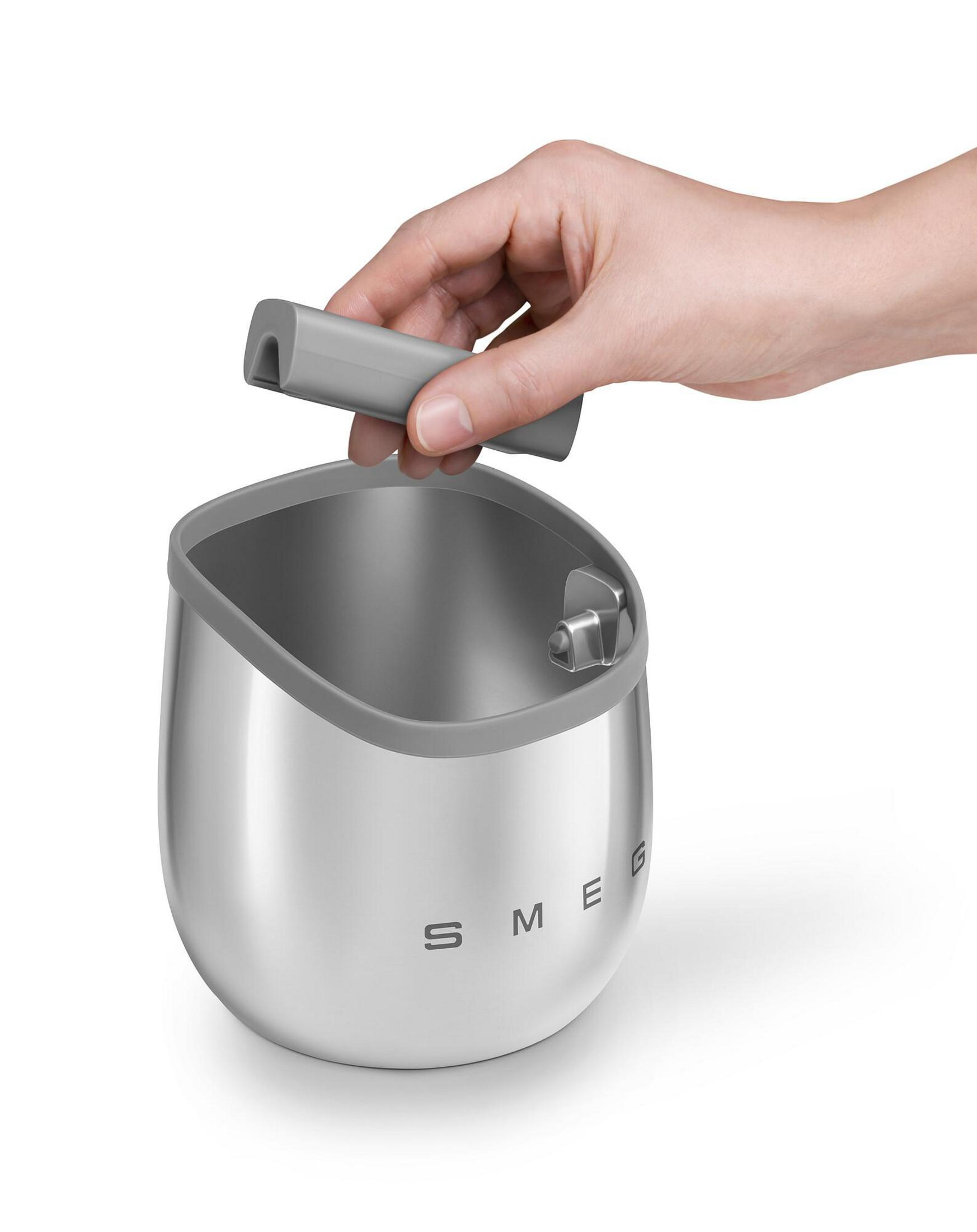 Una mano sostiene una pieza gris sobre un recipiente plateado con la marca SMEG.