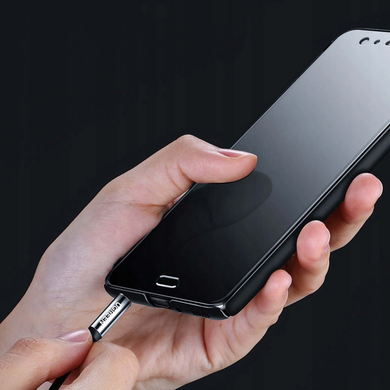 Eine Hand steckt ein silbernes UGREEN-Audiokabel in ein schwarzes Smartphone.
