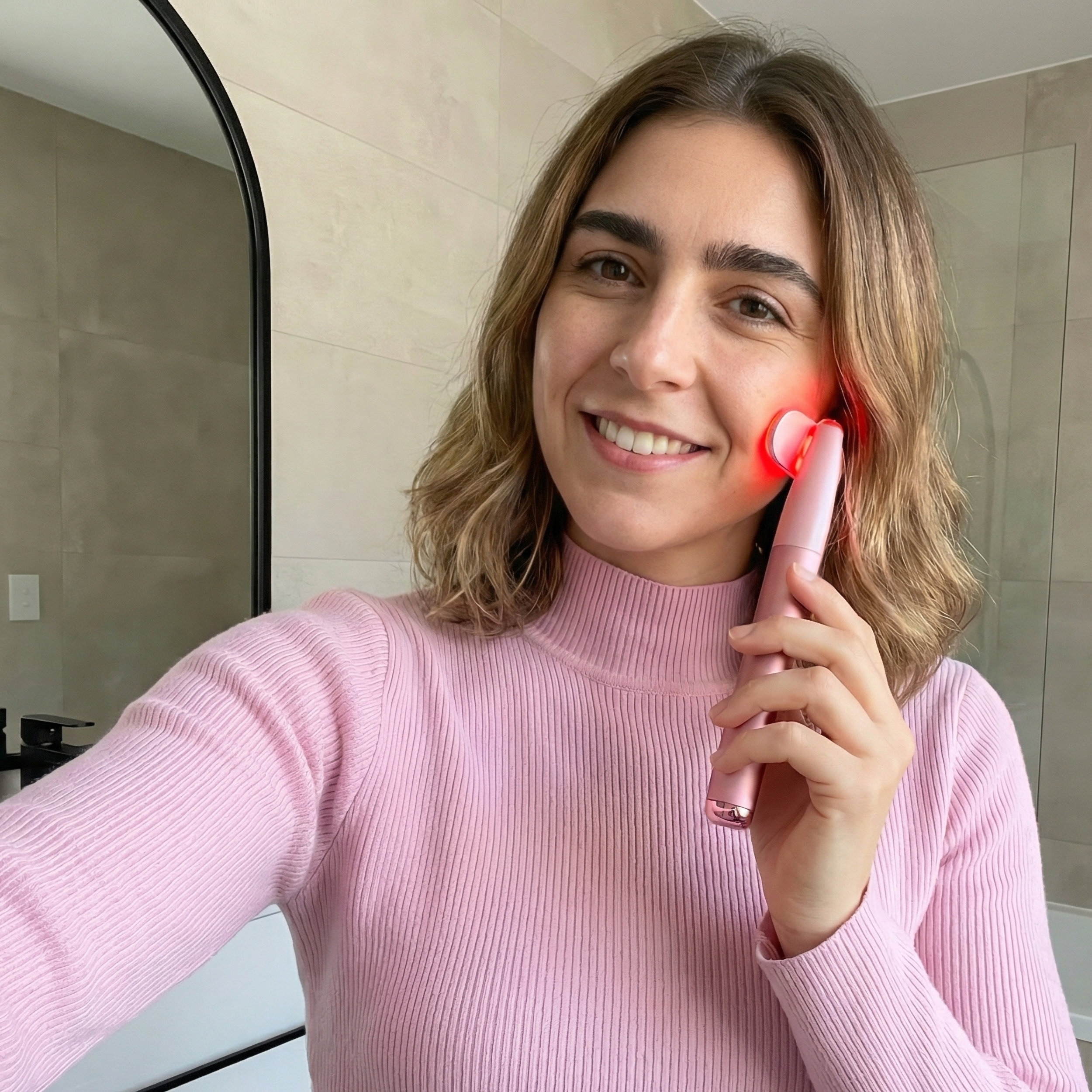 Una mujer con un jersey rosa usa un dispositivo de cuidado de la piel rosa con luz roja.