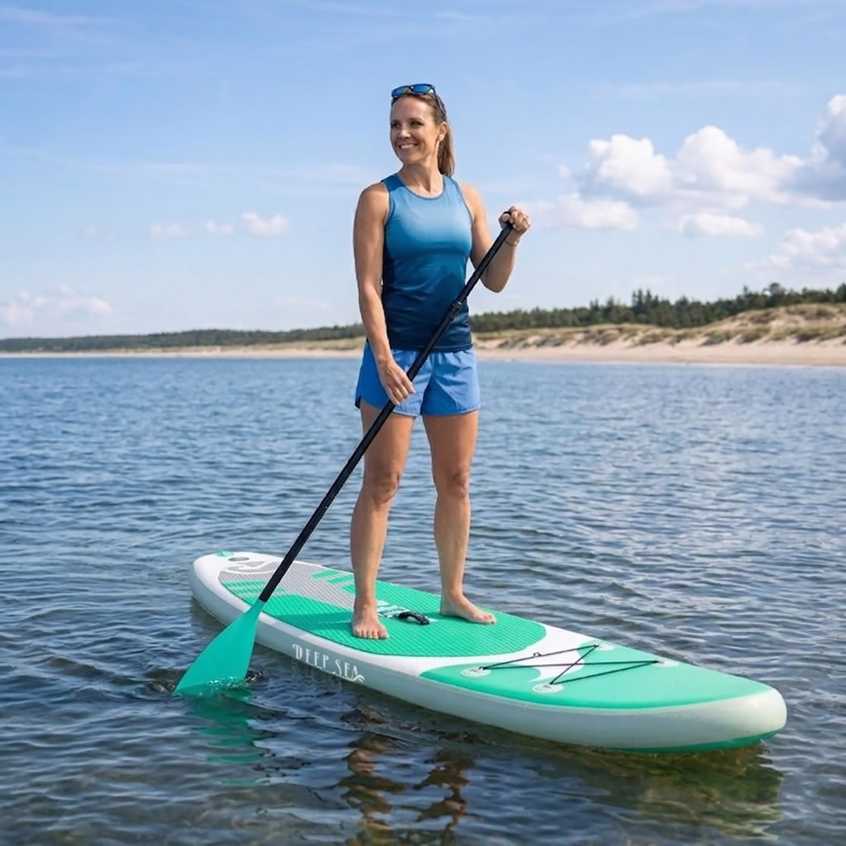 Kobieta pływa na desce z wiosłem po spokojnym jeziorze z piaszczystą plażą.