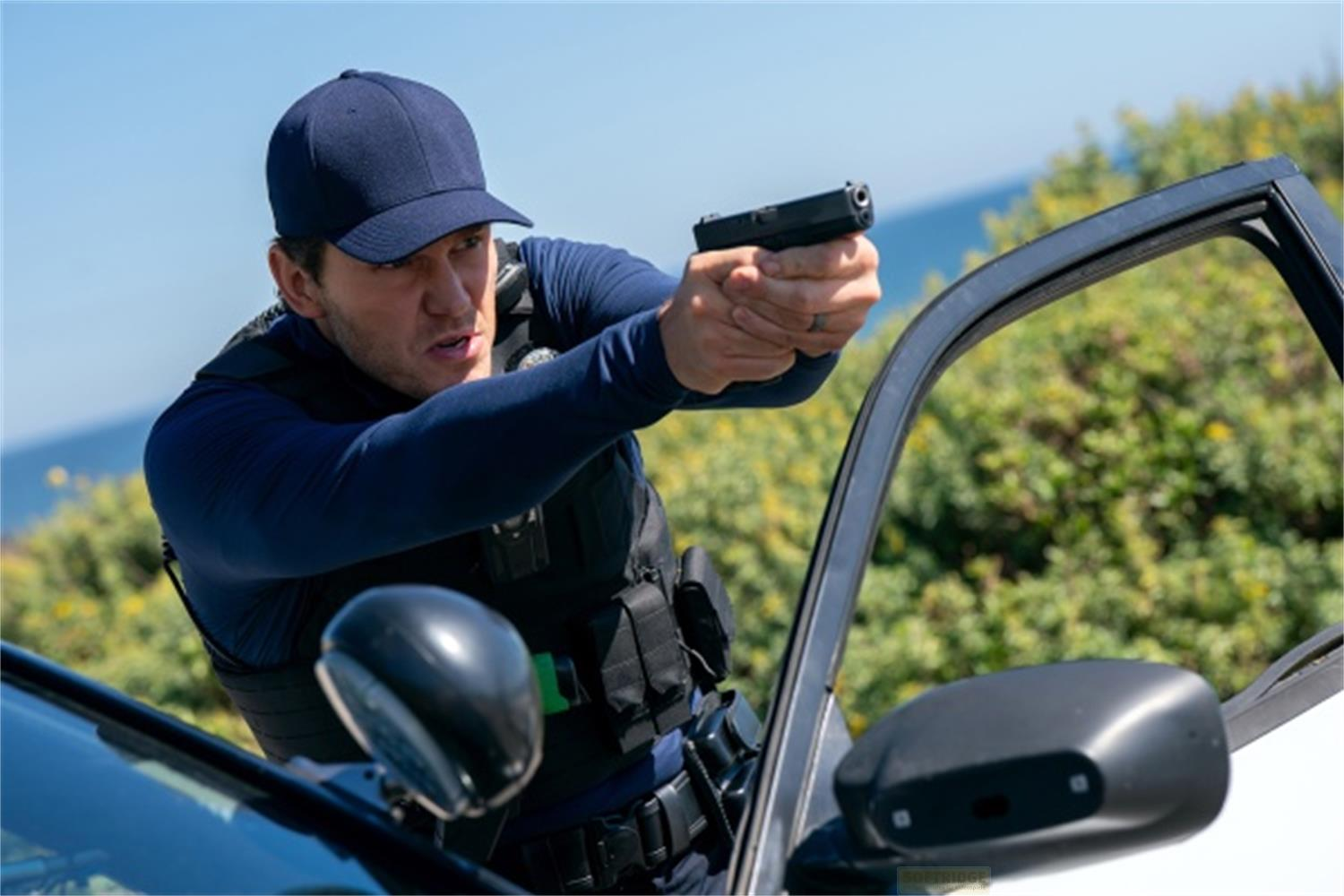 Agente di polizia mira pistola dalla portiera auto, mare blu e cespugli verdi.