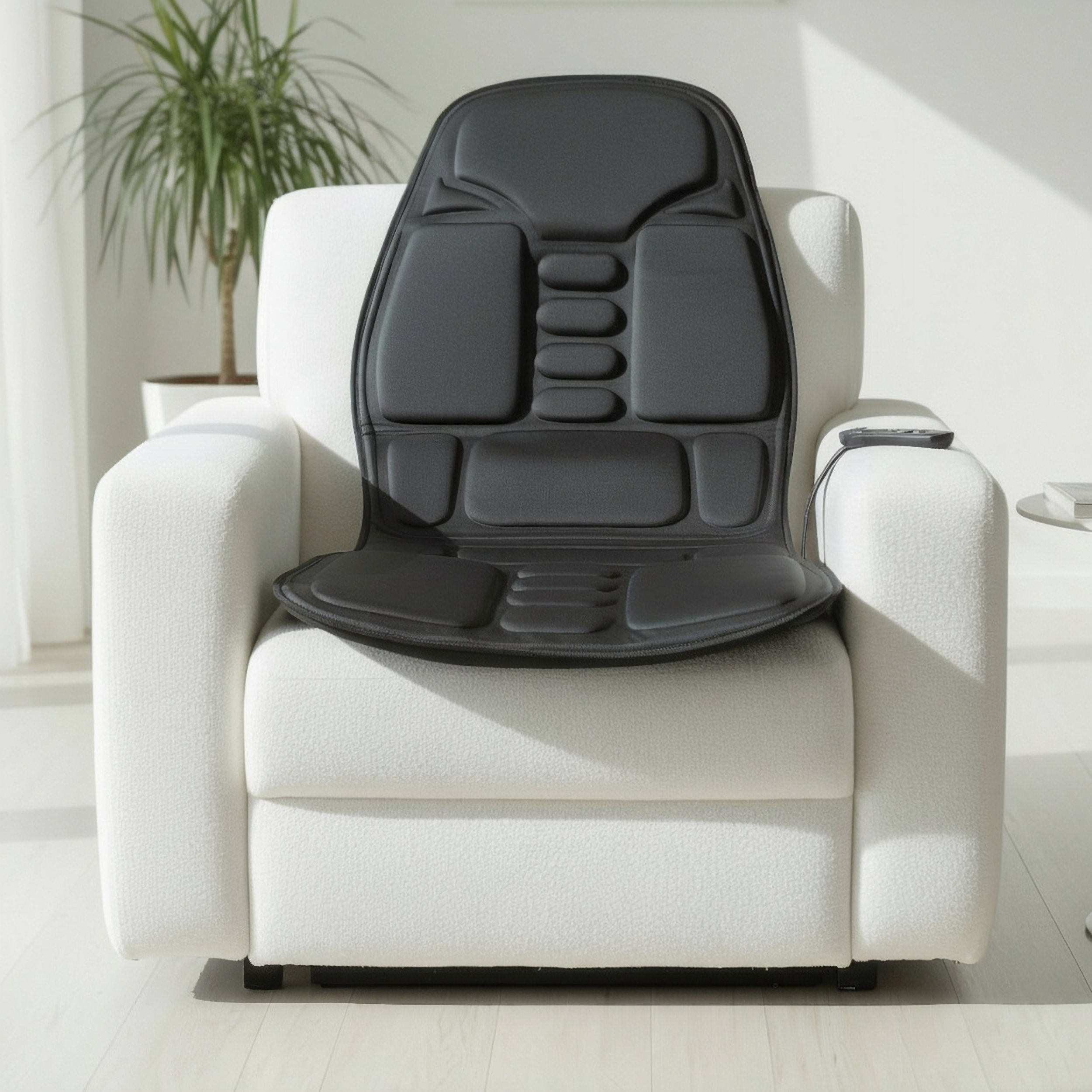 Esterilla de masaje negra sobre un sillón blanco, con mando y planta al fondo.