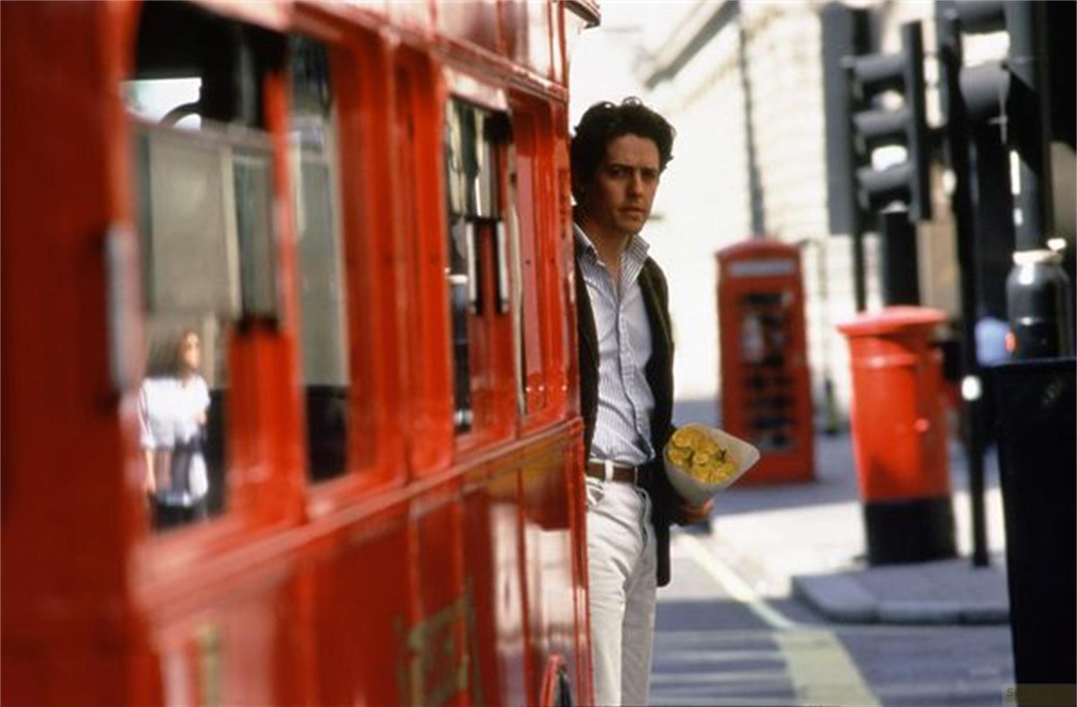 Uomo con mazzo di fiori vicino a bus rosso a due piani, cabina telefonica sullo sfondo.