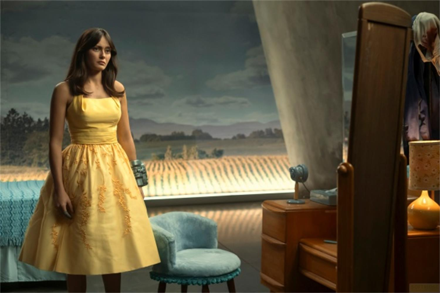 Donna in abito giallo vicino al letto, campi a motivi visibili dalla finestra.