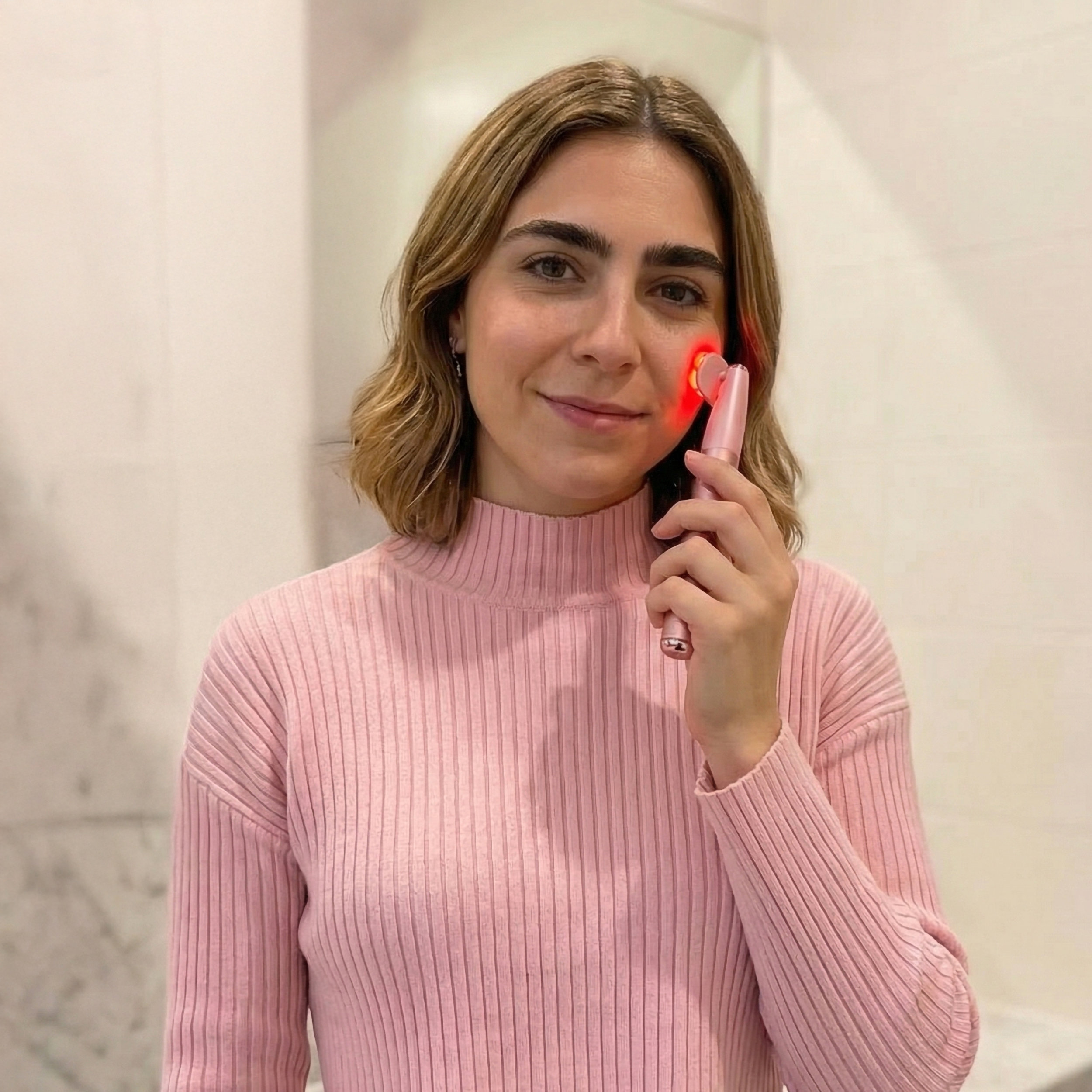 Una mujer con suéter rosa sostiene un dispositivo de cuidado de la piel rosa con luz roja en la cara.
