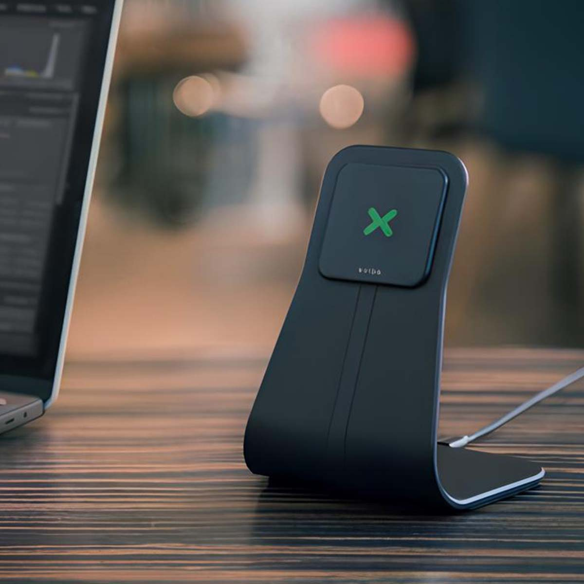 Black wireless charging stand with green X and laptop on wooden desk.