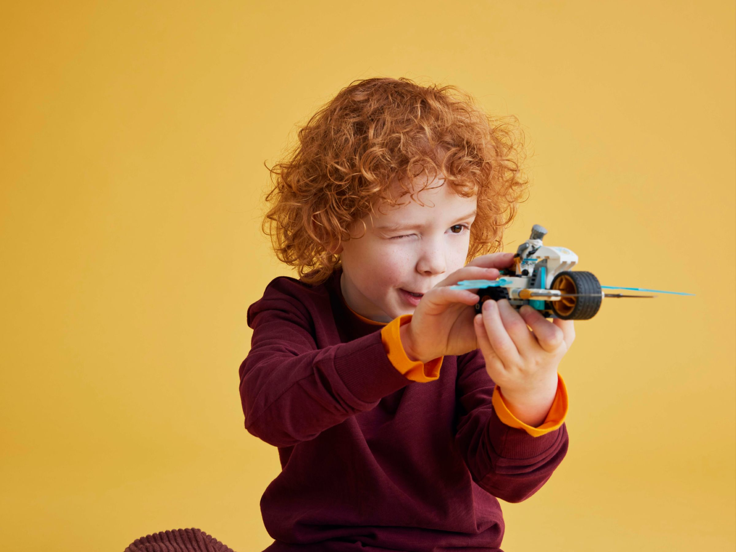 Ein Junge mit roten Locken spielt mit einem Spielzeugauto vor gelbem Hintergrund.