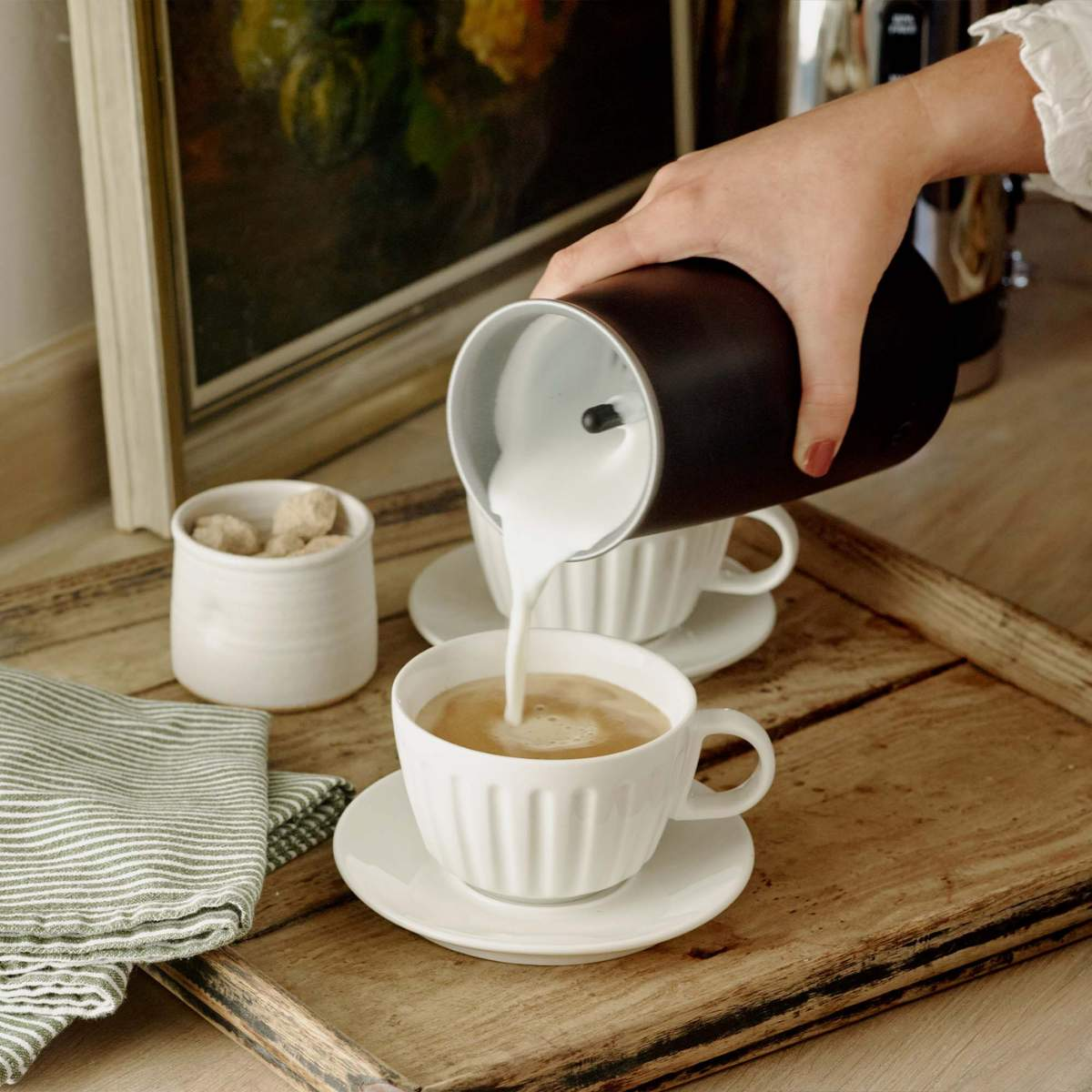 Hand gießt aufgeschäumte Milch in eine Kaffeetasse auf einem Holzteller mit Zuckerwürfeln.