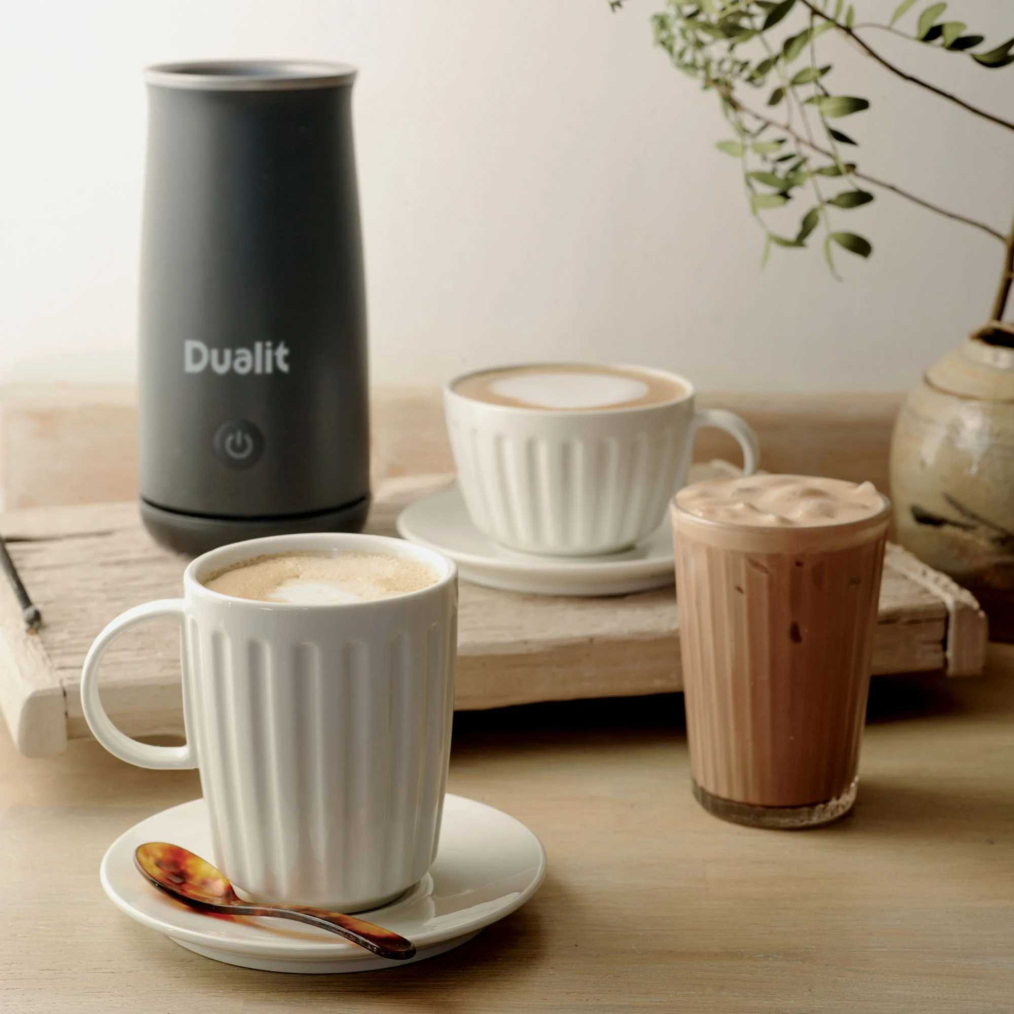 Ein grauer Dualit-Milchaufschäumer neben zwei Tassen Kaffee und einem Glas Schokoladengetränk.