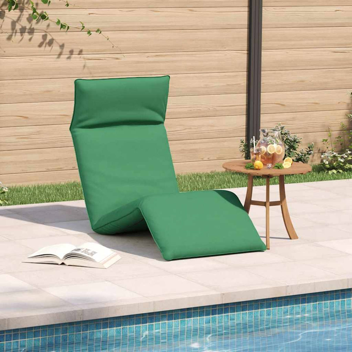 Sdraio verde a bordo piscina con parete in legno, tavolo con caraffa e libro.
