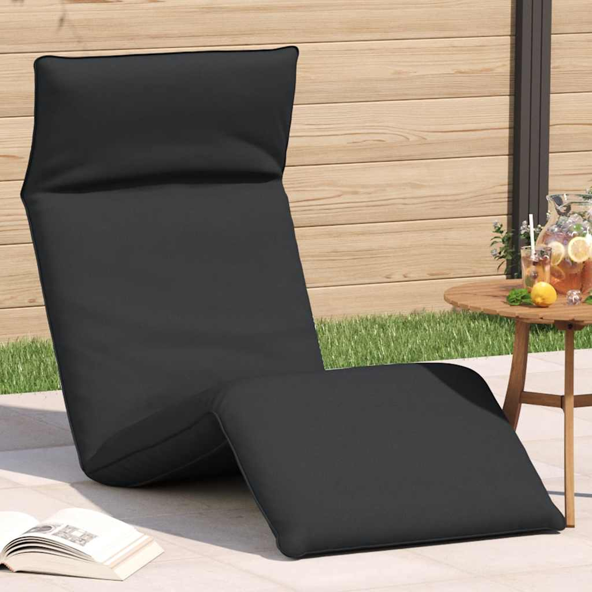 Ein schwarzer Liegestuhl steht auf einer Terrasse neben einem kleinen Tisch mit Getränken und einem Buch.