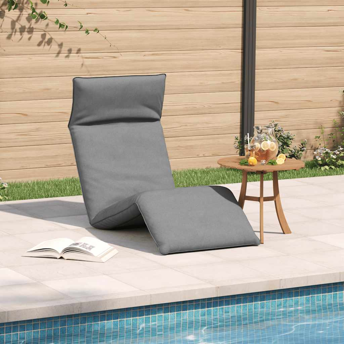 Una poltrona da salotto grigia, un libro e una bevanda su un tavolino di legno vicino a una piscina.