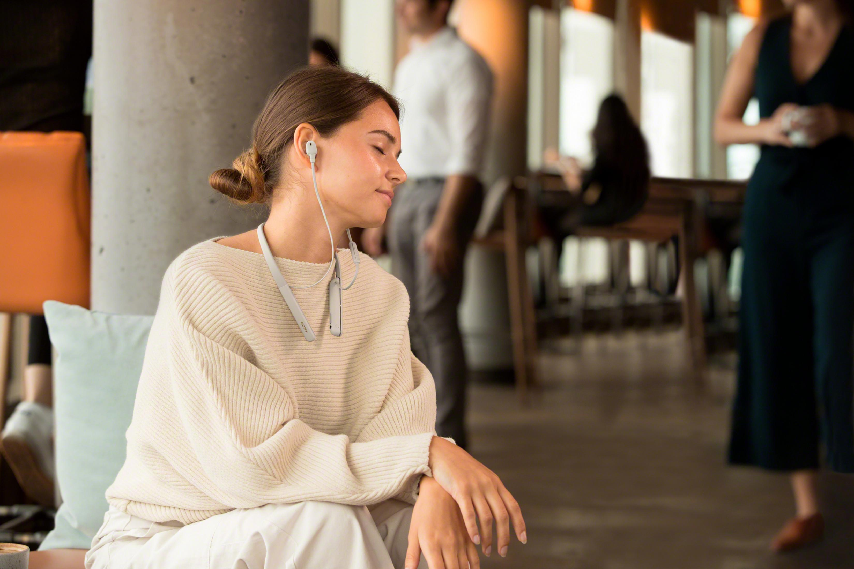 A woman with headphones sits relaxed in a bright room with other people in the background.