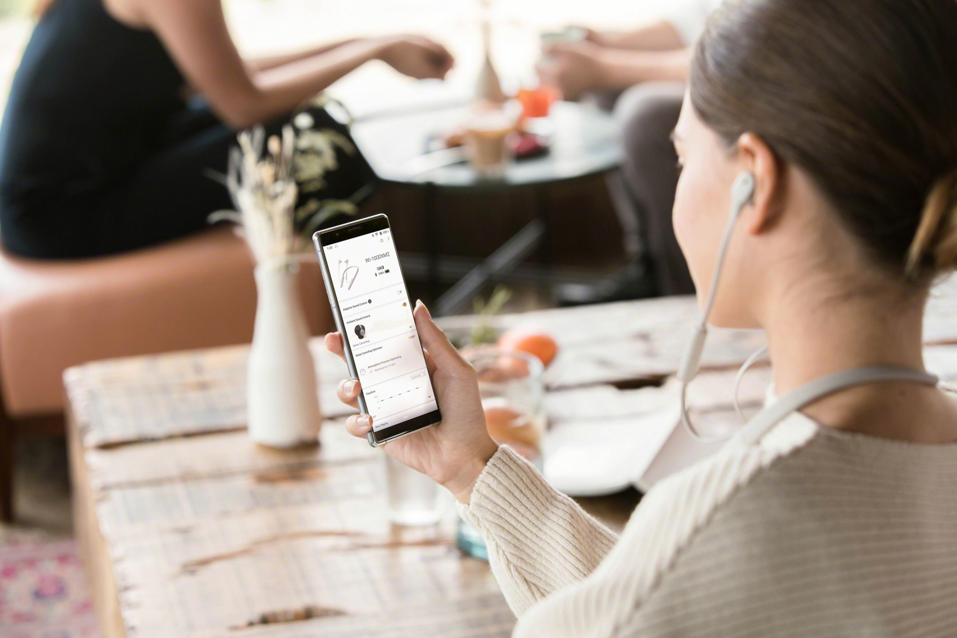 A woman holds a smartphone showing a headphone app. People sit in the background.