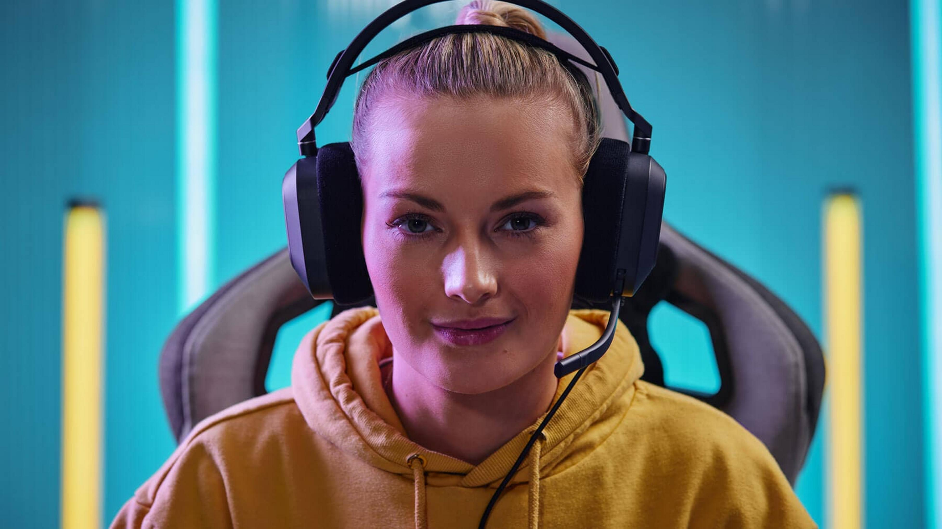 Eine junge Frau mit Kopfhörern und gelbem Hoodie sitzt vor blauem Hintergrund.