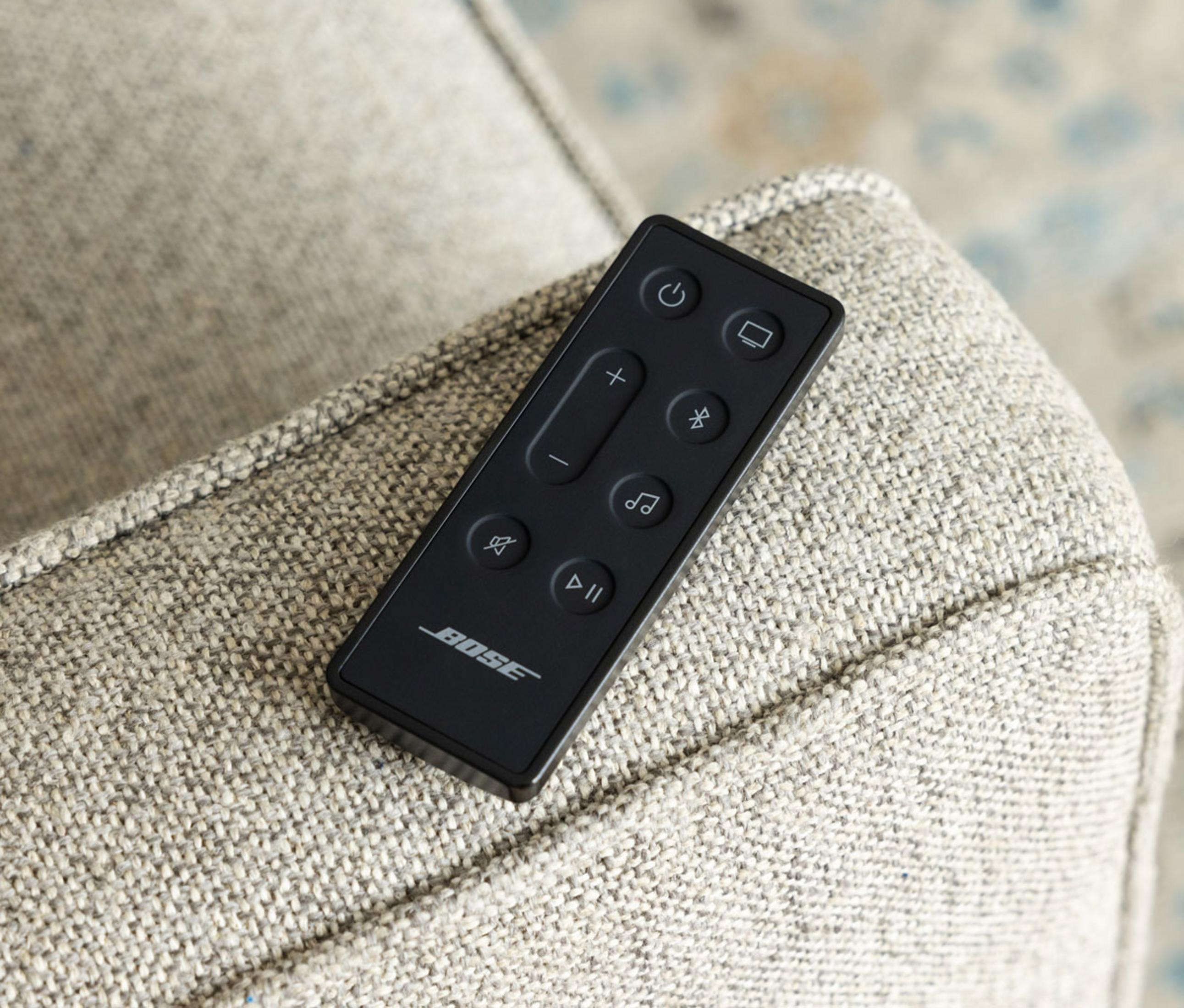 Control remoto Bose negro con botones sobre una silla de tela gris claro.